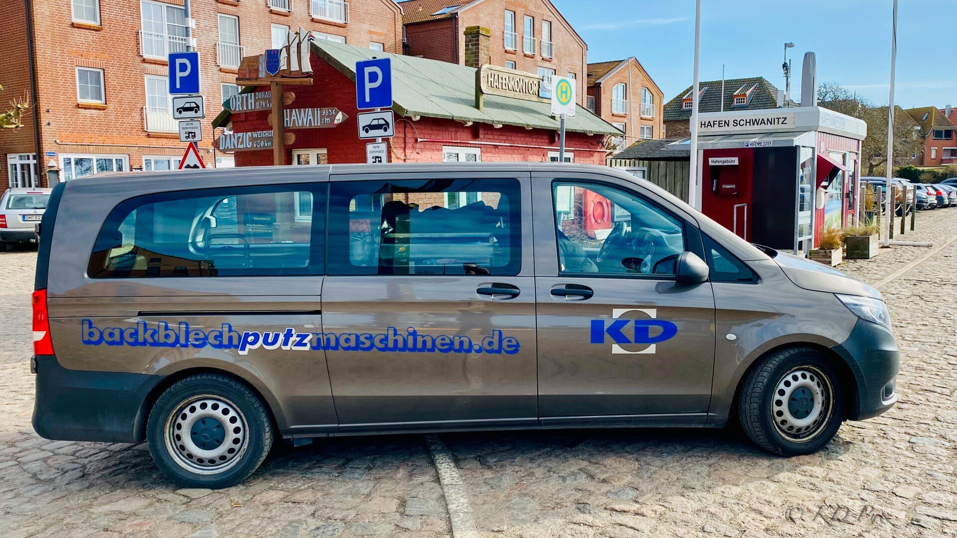 Ein grauer Lieferwagen mit der blauen Aufschrift „backblechputzmaschinen.de“ und „KD“ an der Seite parkt auf einer Kopfsteinpflasterstraße vor Backsteingebäuden und einem roten Kiosk. Zwei blaue Parkschilder sind sichtbar.