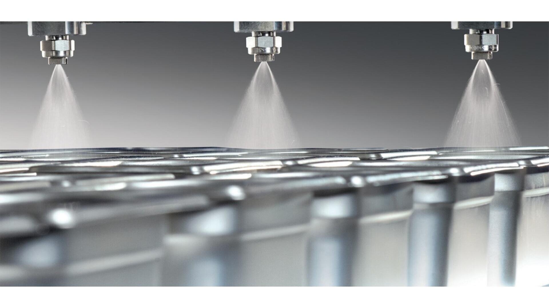 Three spray nozzles evenly coat the tops of metal containers with a fine mist in an industrial setting, suggesting a process like painting, coating, or sanitizing.