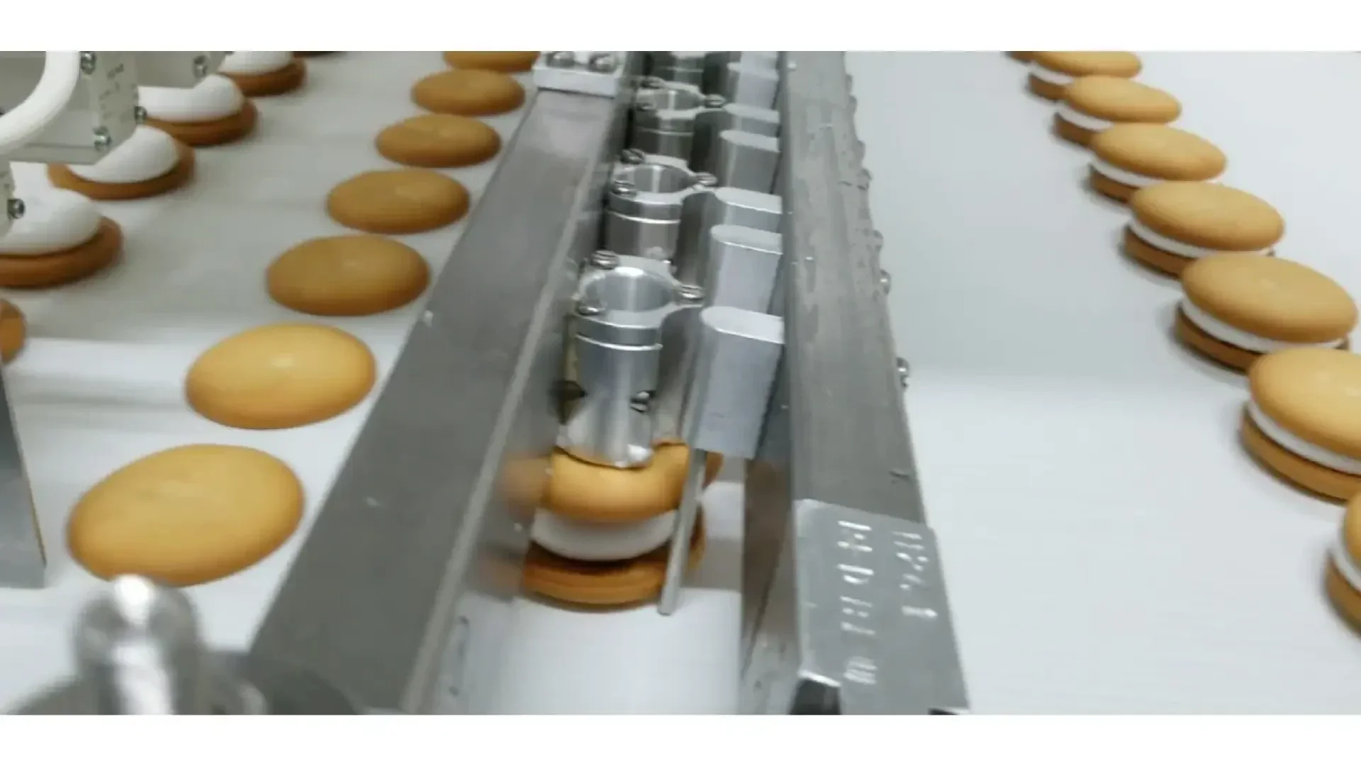 Rows of round cookies with white filling move along a conveyor belt in a factory, as machinery aligns and processes them for packaging.