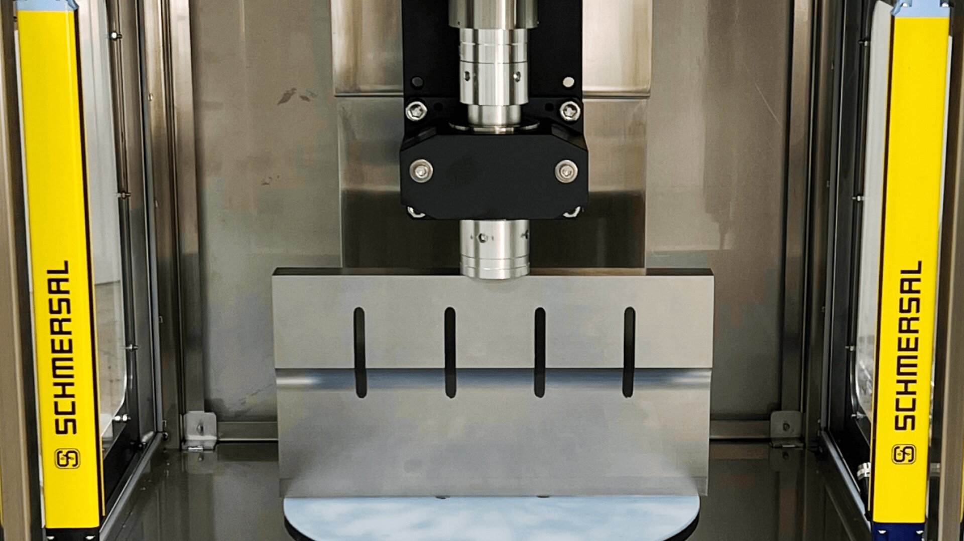 A close-up view of industrial machinery featuring a metal press with a flat plate, safety guards labeled Schmersal on both sides, and a cylindrical component above the press plate.