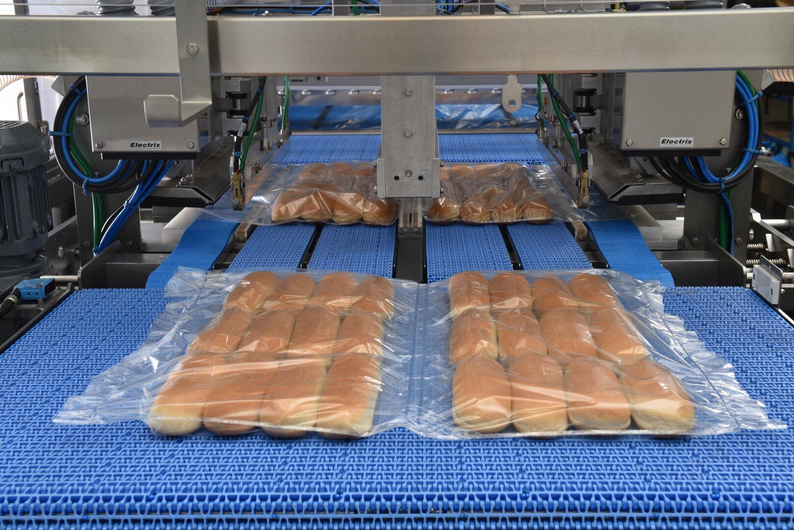 Auf einem blauen Förderband in einer automatisierten Lebensmittelverarbeitungsanlage werden reihenweise Hotdog-Brötchen in Kunststoffverpackungen eingeschweißt. Maschinen umgeben das Band und steuern den Verpackungsprozess.