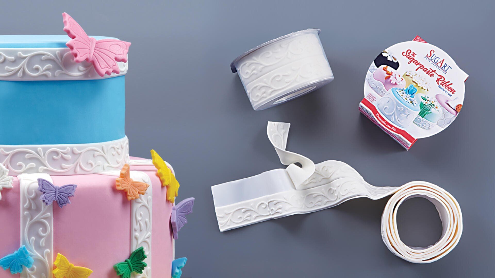 Ein blauer und rosafarbener Stufenkuchen, verziert mit bunten Schmetterlingen und weißem, geprägtem essbarem Band, steht neben einer Rolle und einer Verpackung mit weißem Dekoband aus Zucker auf einer grauen Fläche.