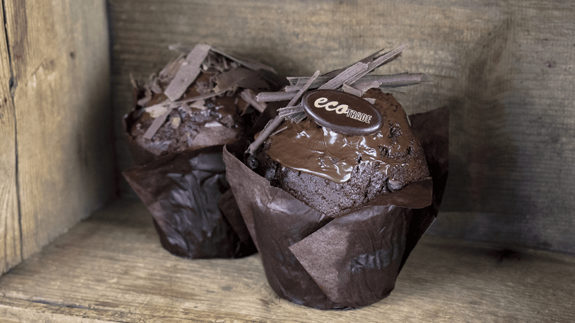 Two chocolate muffins in brown wrappers, topped with chocolate shavings and a small oval chocolate piece with the word eca written on it, set against a rustic wooden background.