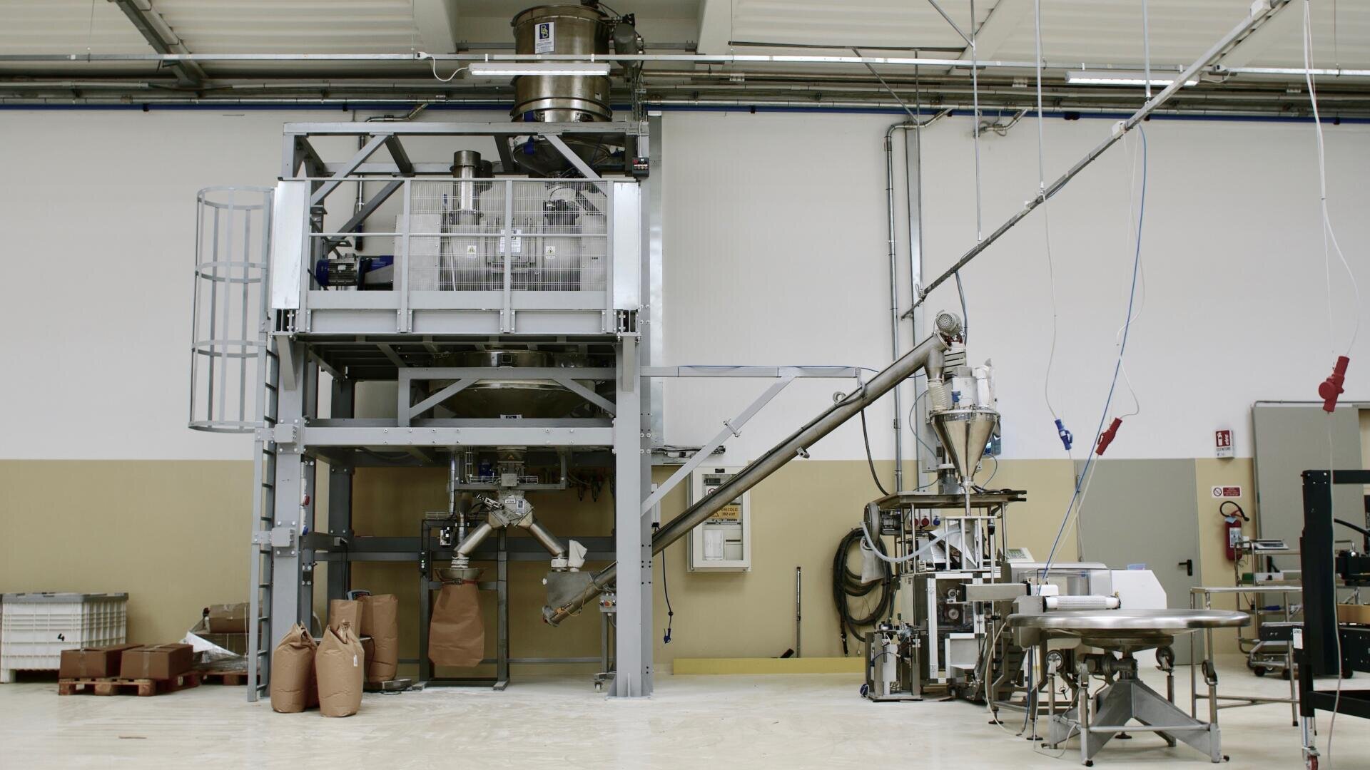 A large industrial packaging machine in a factory, with mechanical arms, conveyor belts, and brown paper bags, surrounded by pipes, cables, and equipment against a beige and white wall.