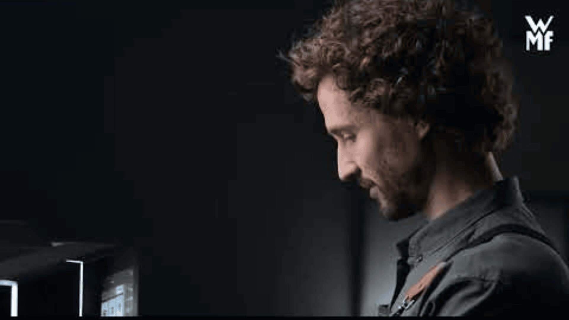 Ein Mann mit lockigem Haar und Bart steht lächelnd neben einer Kaffeemaschine. Der Hintergrund ist dunkel, in der oberen rechten Ecke ist das WMF-Logo zu sehen.