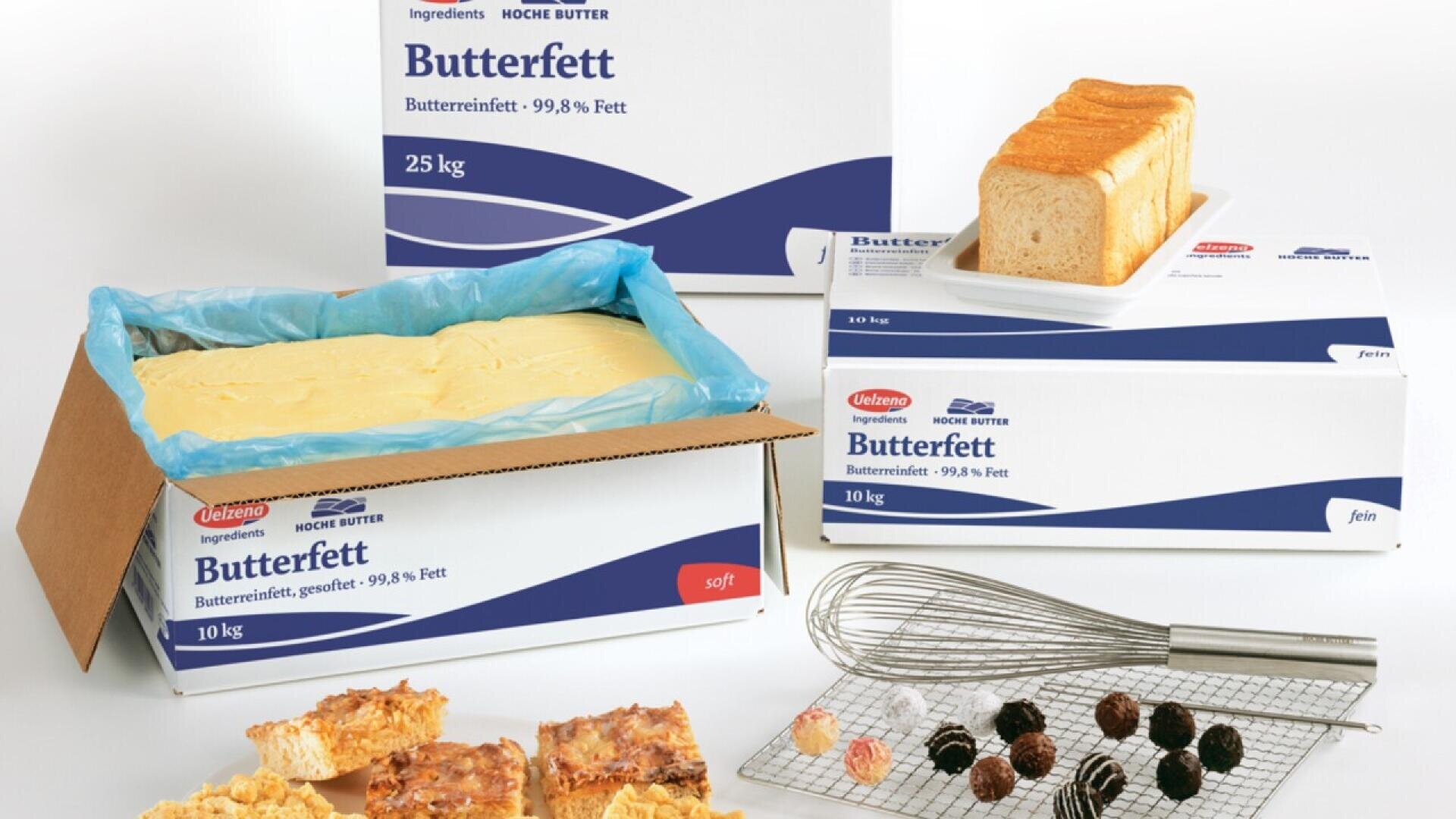 A variety of baked goods, a whisk, and assorted chocolates are displayed in front of large commercial boxes and an open container of Butterfett (butter fat) on a white surface.