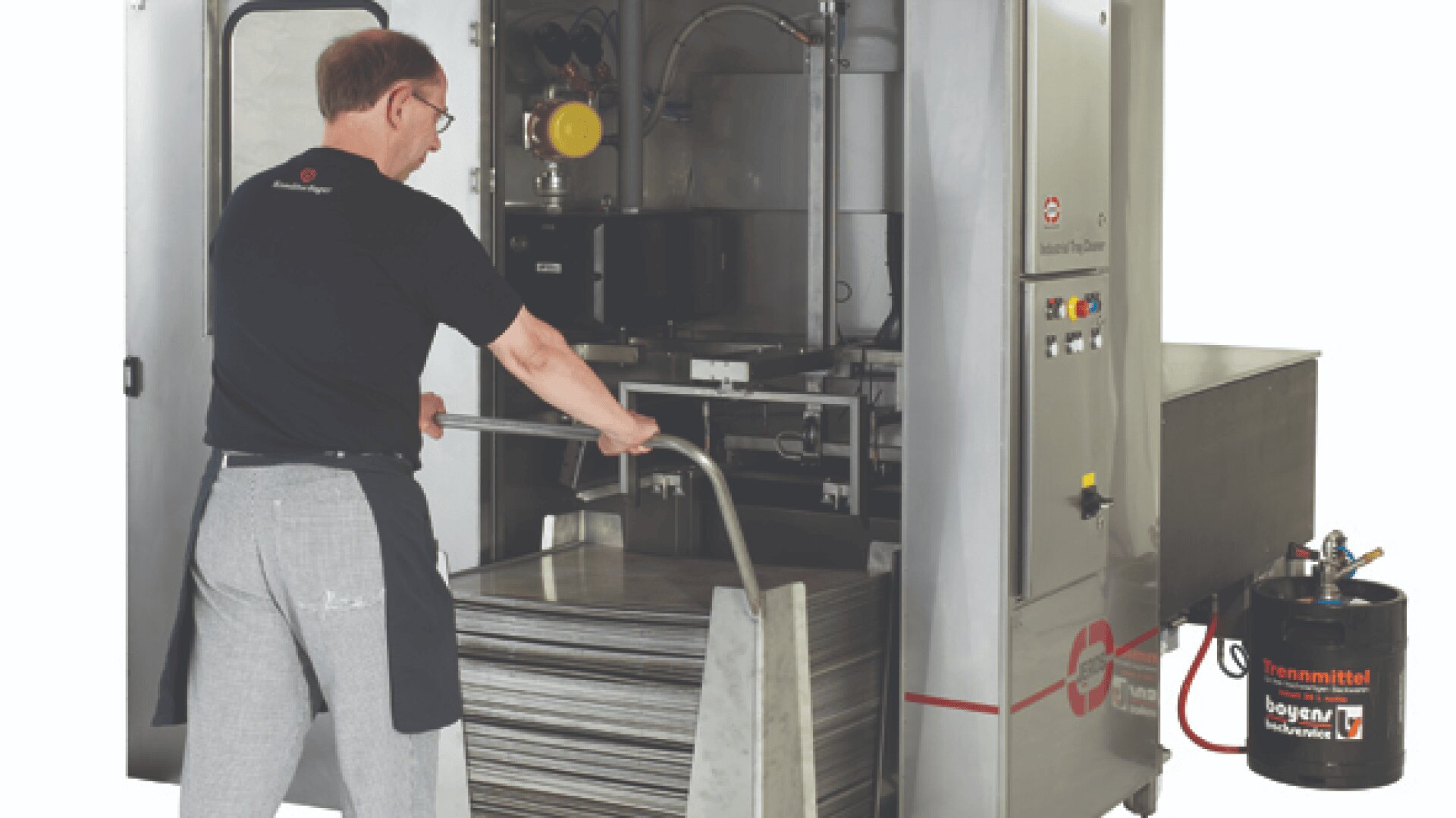 Ein Mann mit Brille und Schürze lädt große Metallschalen in eine industrielle Waschmaschine in einer sauberen, gut beleuchteten Umgebung. Neben der Maschine ist ein Öl- oder Reinigungsmittelbehälter zu sehen.