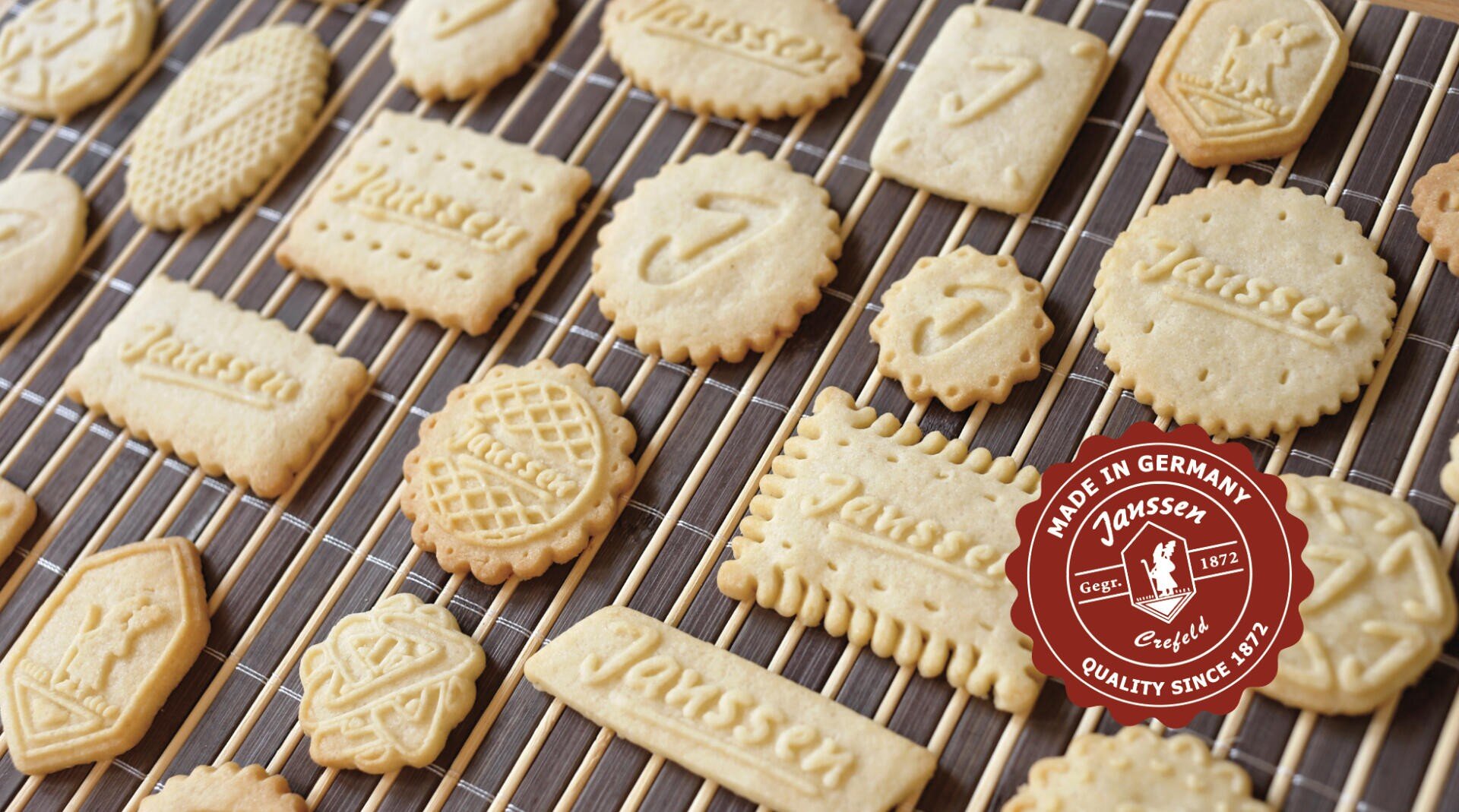 Verschiedene geprägte Butterkekse, die auf einem Drahtgestell abkühlen. Die Kekse haben verschiedene Formen und Muster, einige sind mit "Janssen" beschriftet. In der unteren rechten Ecke befindet sich ein rotes Siegel "Made in Germany, Janssen".