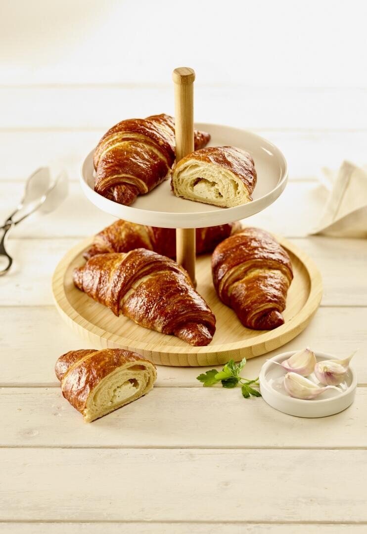 Auf einem zweistöckigen Ständer stehen goldbraune Croissants, ein Croissant ist aufgeschnitten, damit man seine flockigen Schichten sehen kann. Auf dem Tisch stehen eine Zange, eine Stoffserviette und eine kleine Schale mit Knoblauchzwiebeln, alles auf einer hellen Holzfläche.
