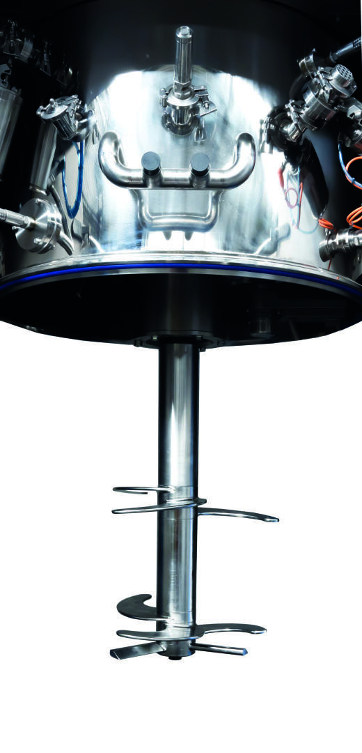 A close-up view of a large industrial stainless steel mixing tank with a central agitator shaft and multiple blades, used for blending or processing materials in manufacturing or laboratory settings.