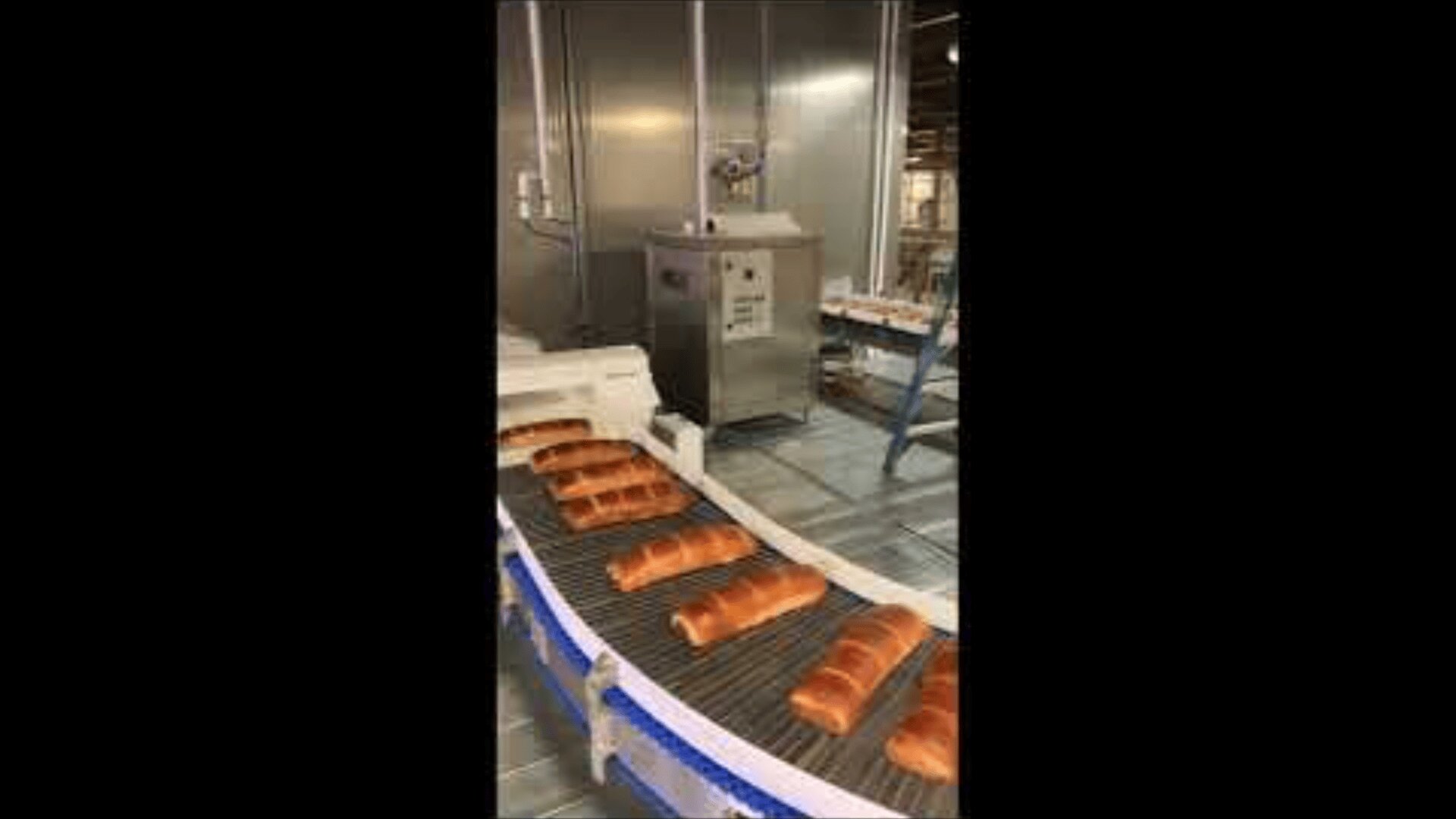 Loaves of bread move along a conveyor belt in a large industrial bakery with metal equipment and machinery in the background.