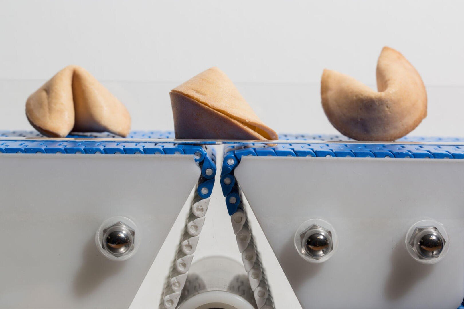 Drei Glückskekse befinden sich nebeneinander auf einem blau-weißen Förderband mit weißem Hintergrund. Die Kekse befinden sich in verschiedenen Stadien des Faltens.