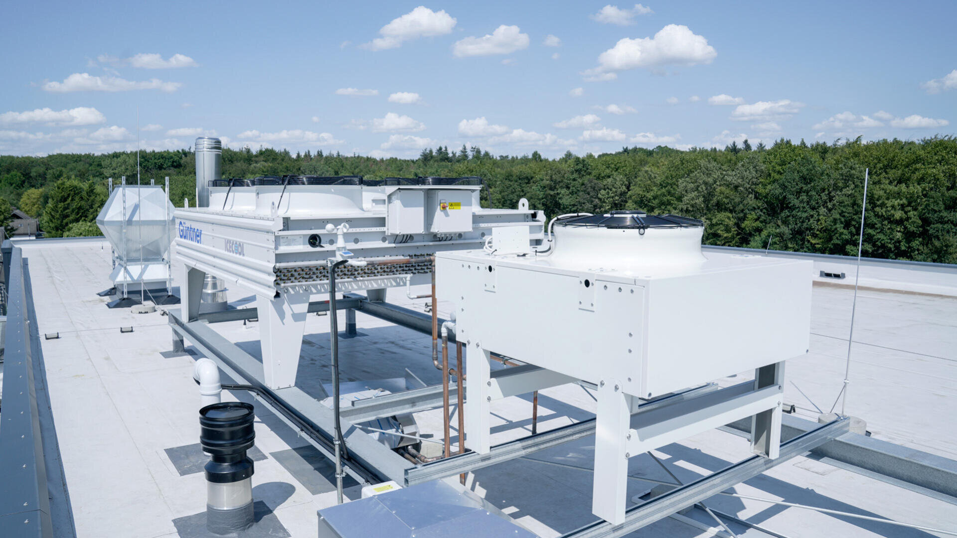Industrielle HLK-Anlagen und Lüftungsgeräte auf einem flachen Dach, umgeben von einem Wald aus grünen Bäumen unter einem blauen Himmel mit vereinzelten Wolken.