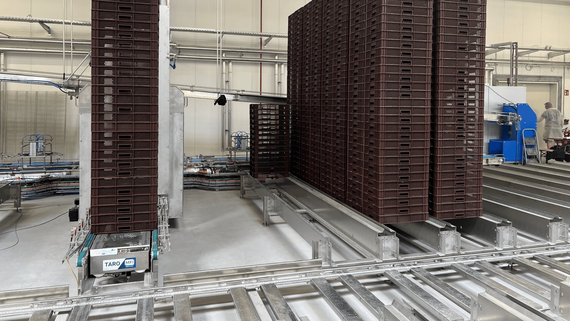 In einer Fabrik sind hohe Stapel brauner Kunststoffkisten auf automatischen Förderbändern zu sehen, in der Nähe befinden sich Industrieanlagen und ein TARO AGV-Roboter. Zwei Arbeiter sind im Hintergrund an blauen Maschinen zu sehen.