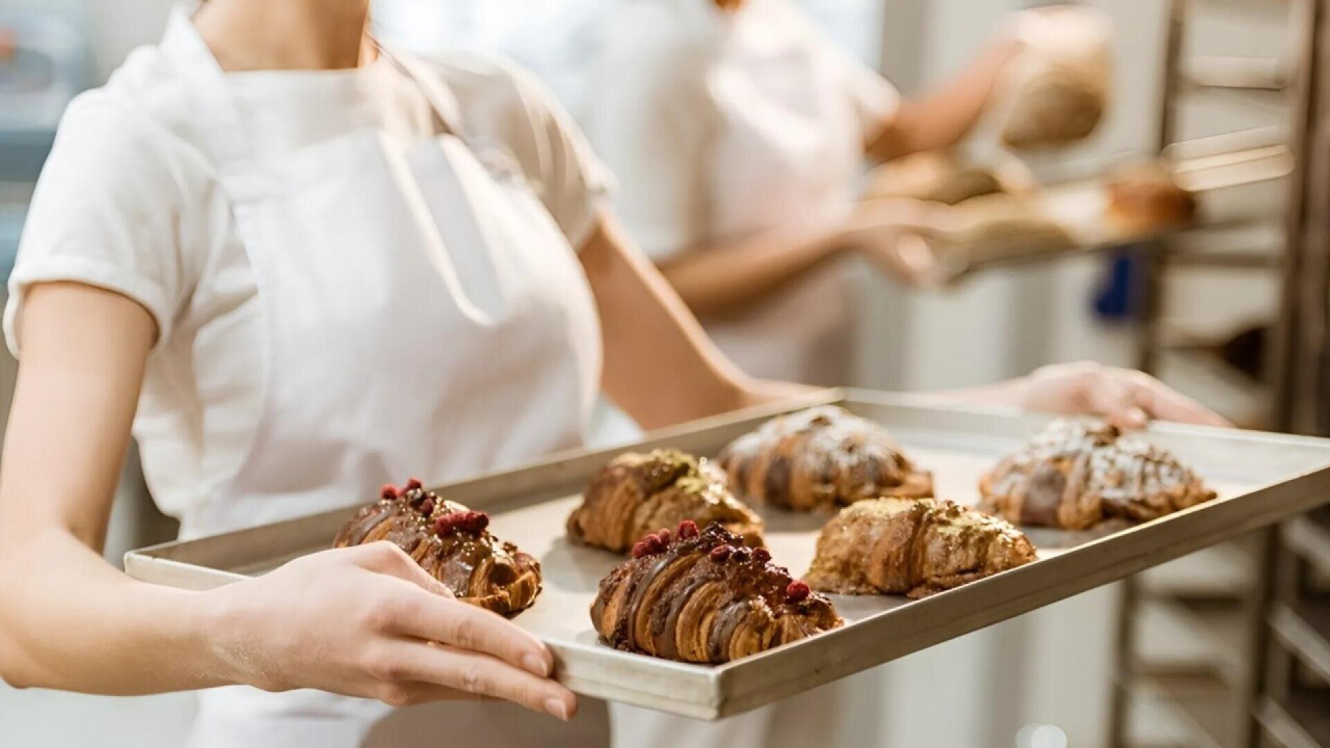 A person wearing a white apron holds a tray of fresh croissants topped with nuts and berries in a bakery, with another person in the background holding a similar tray.