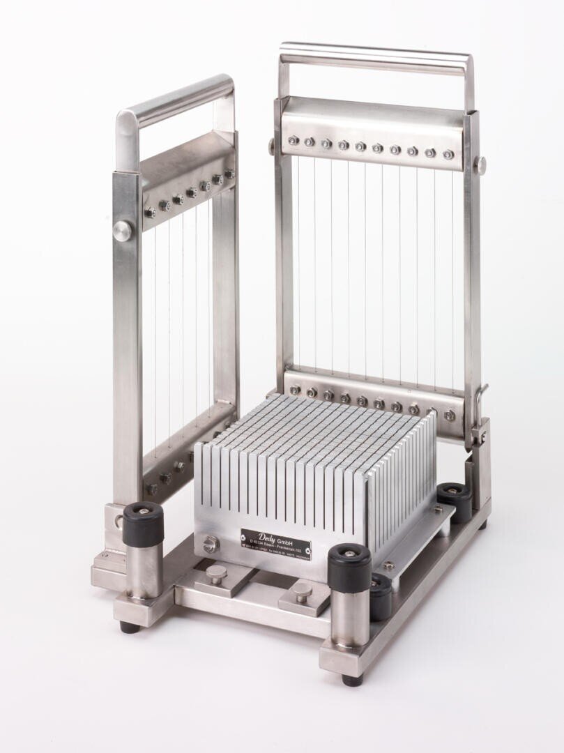 Ein Gerät aus rostfreiem Stahl mit parallelen Drähten und einem beweglichen Rahmen, das zum Schneiden oder Portionieren von weichem Material bestimmt ist. Der zentrale Block scheint für präzise, gleichmäßige Schnitte eingestellt zu sein. Der Hintergrund ist einfarbig weiß.