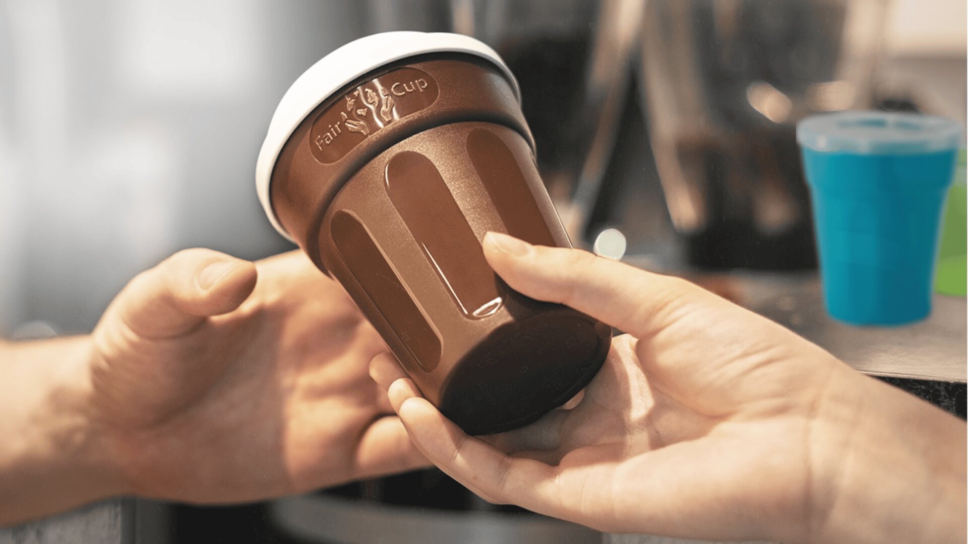 Eine Person reicht einer anderen Person einen braunen Mehrweg-Kaffeebecher mit weißem Deckel, wobei beide den Becher vorsichtig halten. Der Fokus liegt auf den Händen und der Tasse, mit einem unscharfen Hintergrund eines Cafés oder einer Kaffeestation.