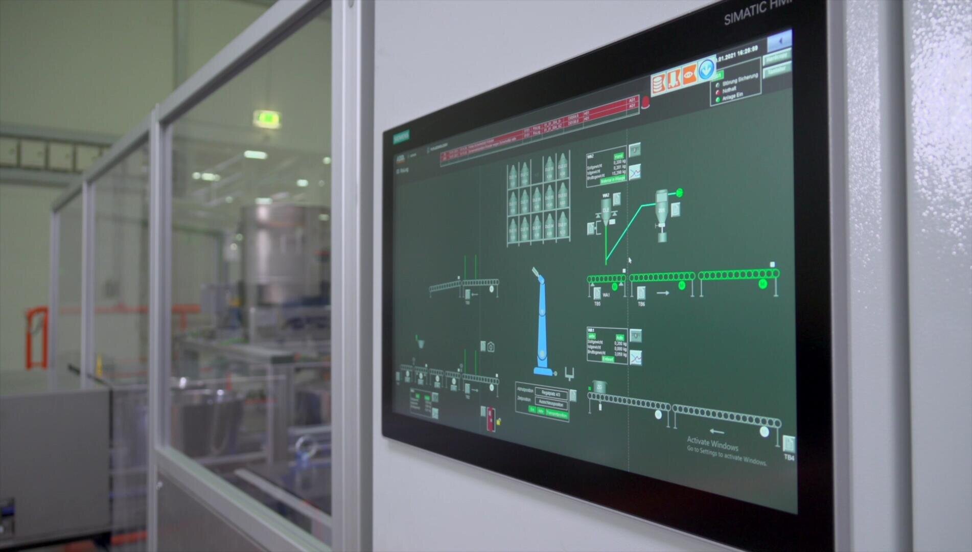 A close-up of an industrial control panel screen displaying a schematic diagram and system status in a factory or manufacturing setting, with machinery visible in the background through glass panels.