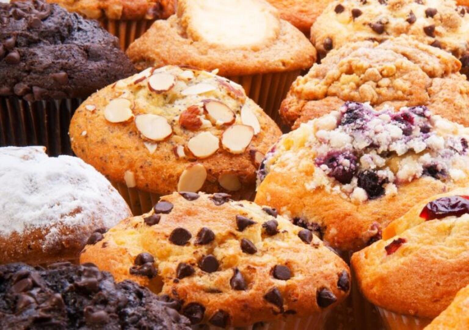 An assortment of muffins with various toppings, including chocolate chips, sliced almonds, powdered sugar, blueberries, and a crumbly streusel, all arranged closely together.