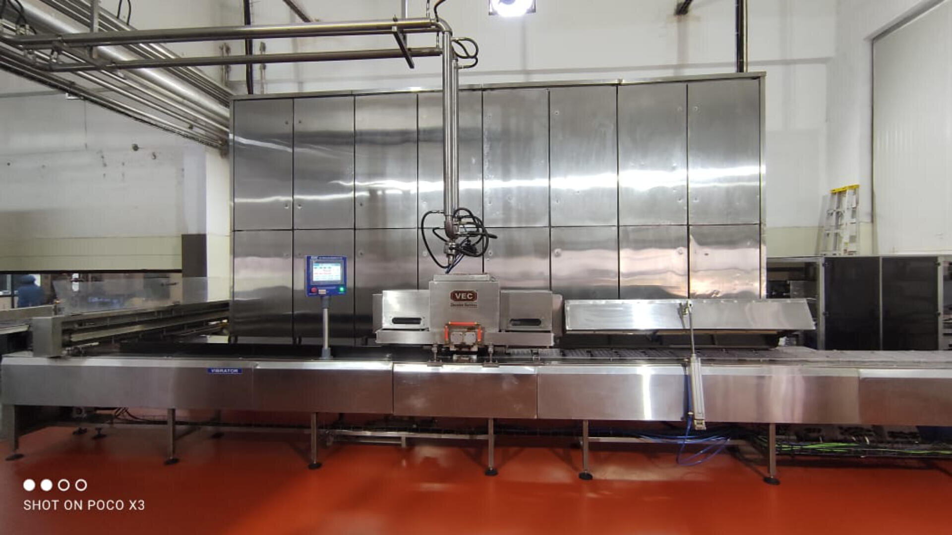 A large stainless steel industrial food processing machine with a conveyor belt stands in a clean, bright factory setting. Pipes run overhead and a digital control panel is attached to the machine. The floor is red.