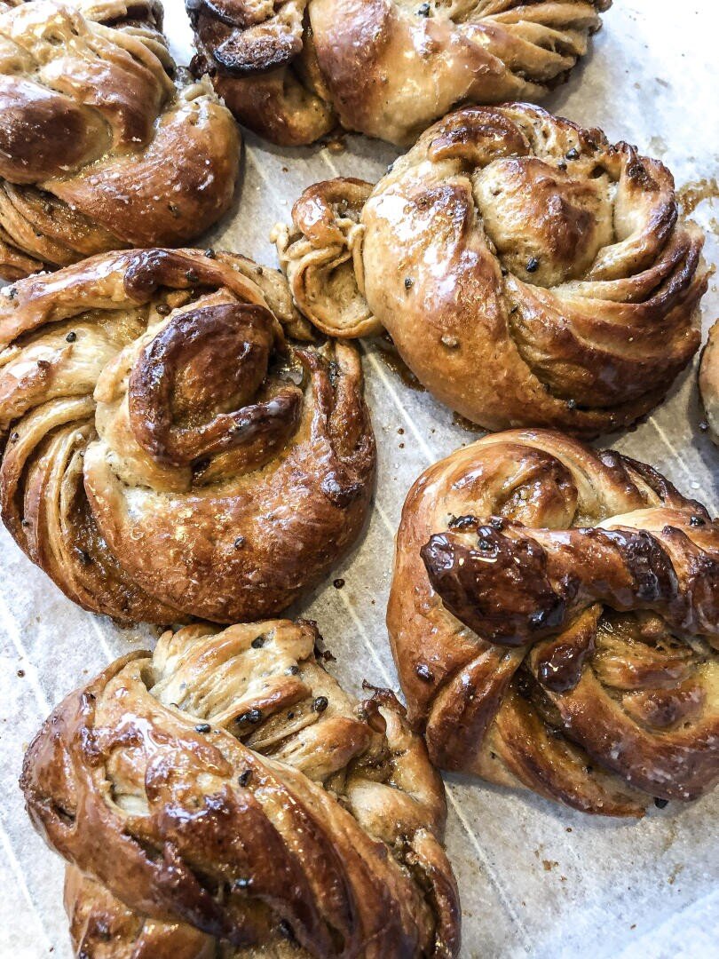Several golden brown, twisted cardamom buns with a glossy finish rest on a sheet of parchment paper. The buns appear flaky and freshly baked, with visible swirls and scattered cardamom seeds.