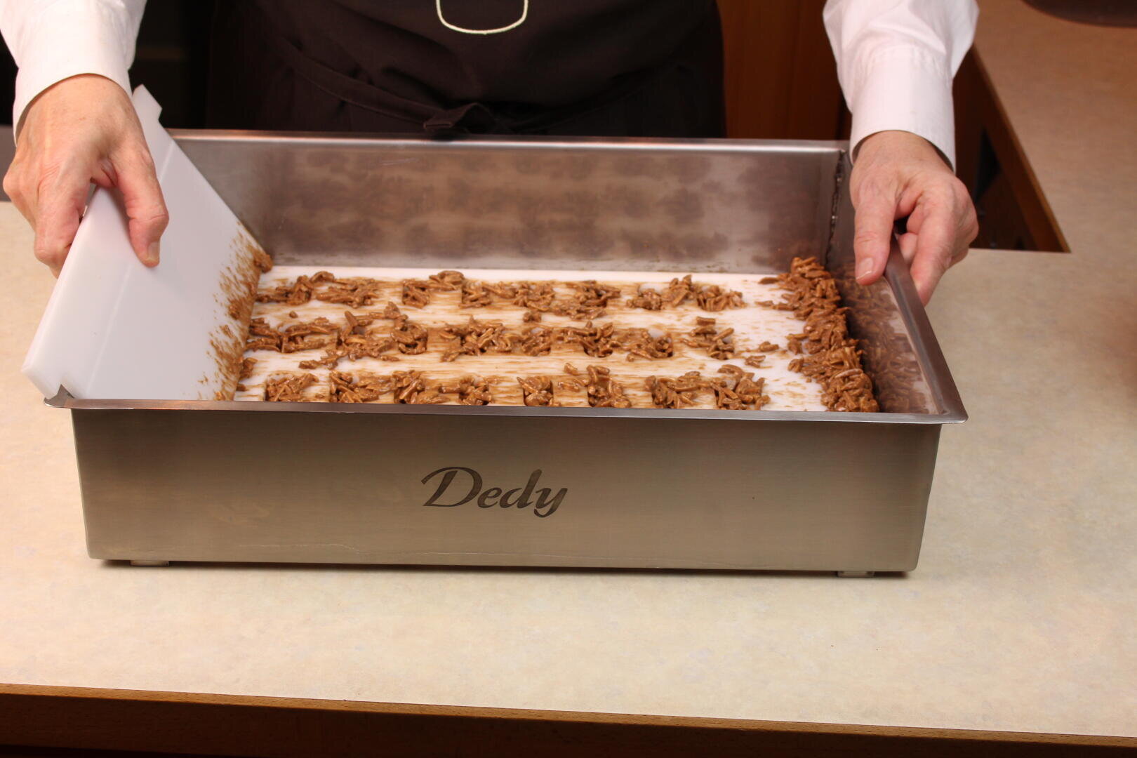 Eine Person verteilt mit einem weißen Spachtel reihenweise Schokolade oder Nougat auf einem großen rechteckigen Metalltablett mit der Aufschrift Dedy, das auf einer Küchenzeile steht.