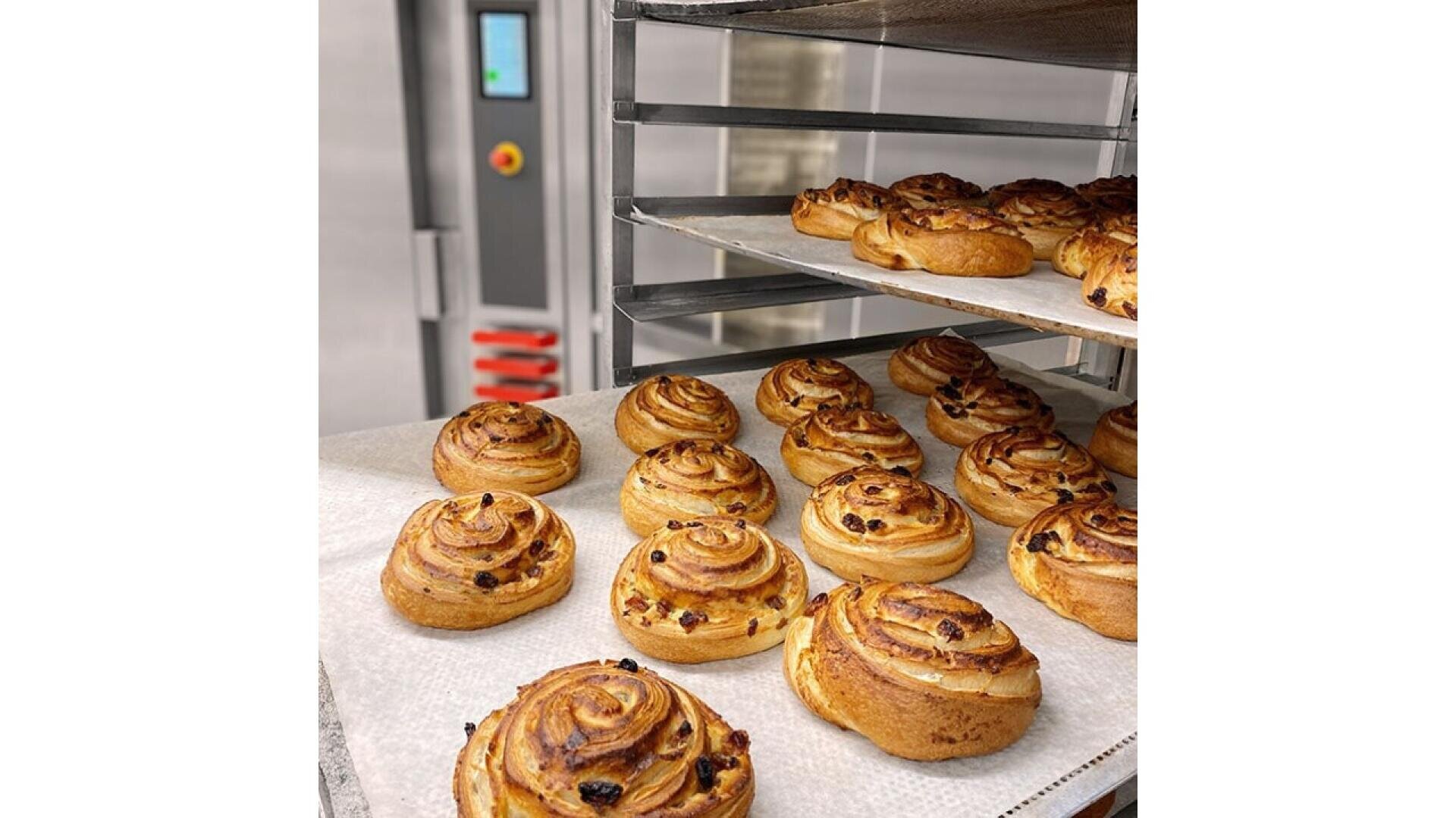 Golden brown cinnamon rolls with raisins are cooling on trays in a commercial bakery. Some trays are on a metal rack, and a large industrial oven is visible in the background.