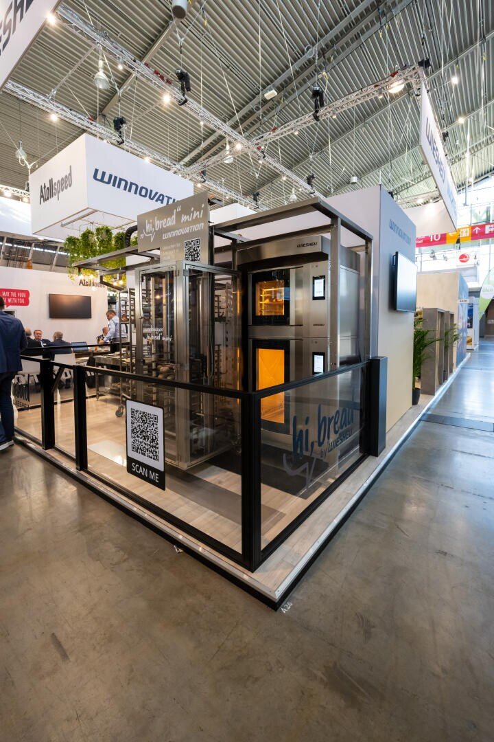 Ein modernes kommerzielles Brotbackofen-Display auf einer Messe, umgeben von Glasscheiben mit QR-Codes. Auf den Schildern steht hi.bread mini und Winnowate. Der Bereich ist gut beleuchtet, mit sichtbaren Messeständen und Besuchern im Hintergrund.