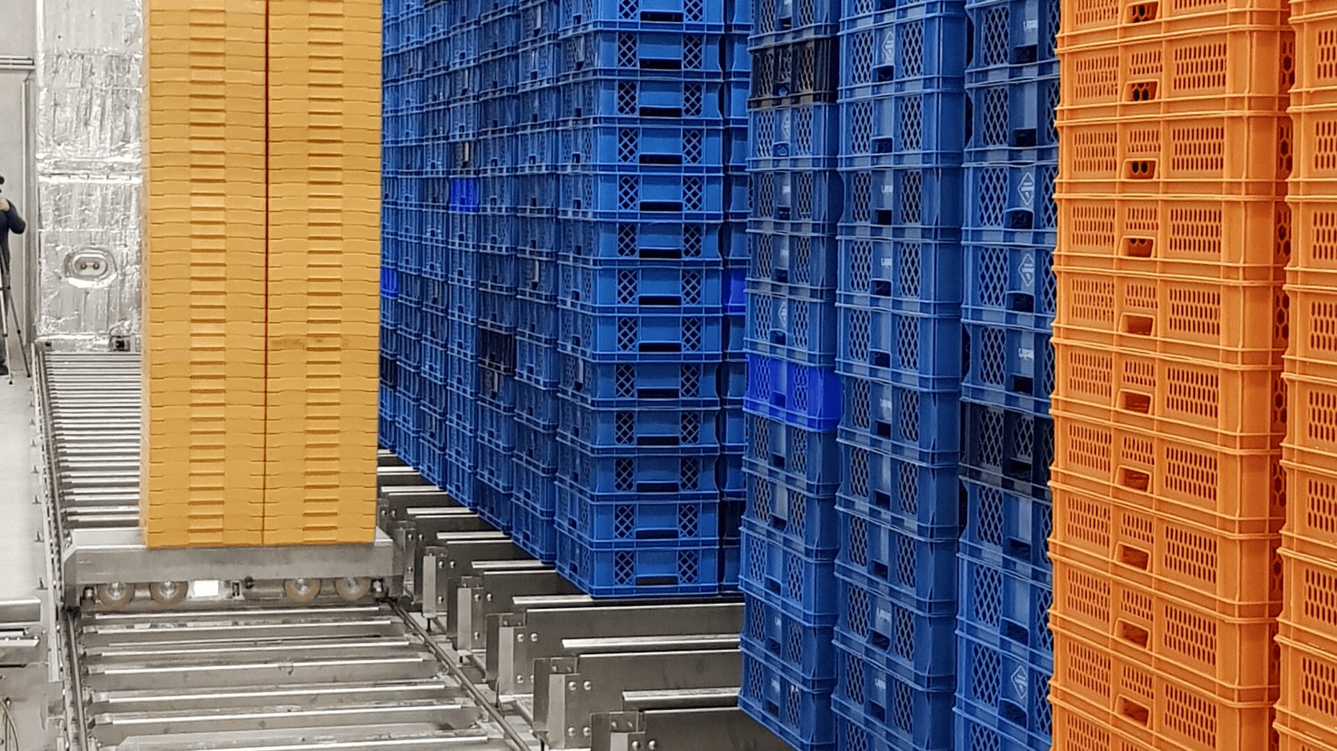 Stapel von gelben, blauen und orangefarbenen Kunststoffkisten sind auf Metallrollen in einer industriellen Lagerumgebung angeordnet. Die Kisten bilden hohe, geordnete Säulen.