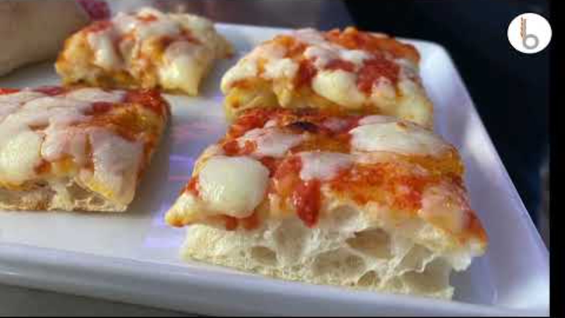 Nahaufnahme von dicken, rechteckigen Pizzascheiben mit geschmolzenem Käse und Tomatensauce auf einem weißen Teller. Die Pizza hat eine leichte, luftige Kruste mit sichtbaren Blasen.