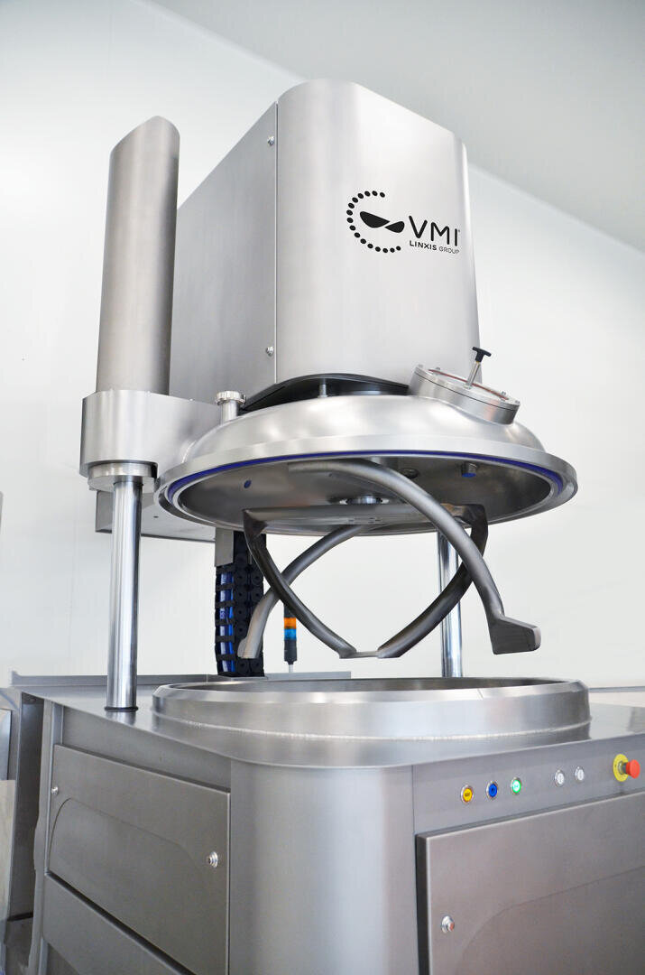 Large industrial stainless steel mixer with a VMI logo, featuring mixing arms and a circular bowl below, situated in a clean, modern facility. Control buttons are visible on the front panel.