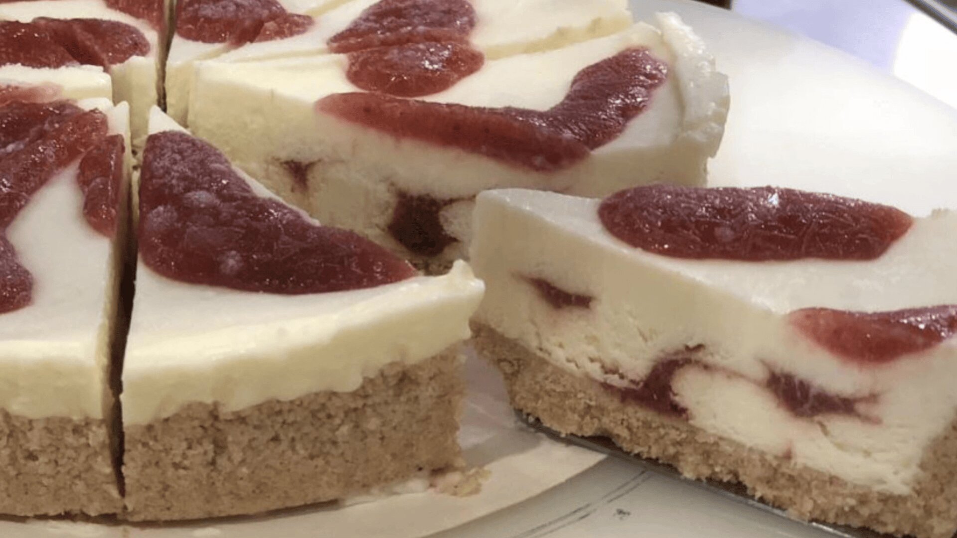 Close-up of sliced cheesecake with a thick graham cracker crust and swirls of red strawberry sauce throughout the creamy filling and on top.
