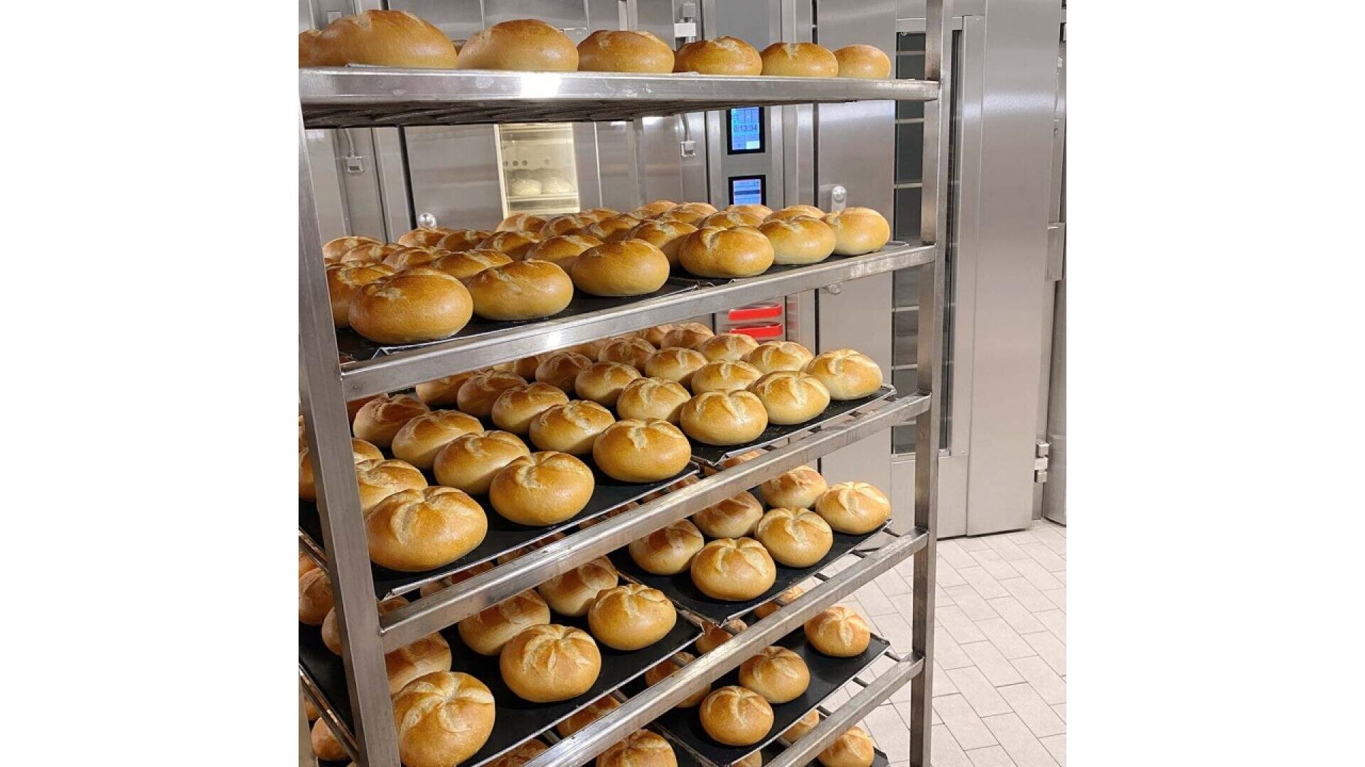 Metal baking racks filled with rows of round, golden-brown bread rolls in a commercial kitchen or bakery with stainless steel equipment and tiled floors in the background.