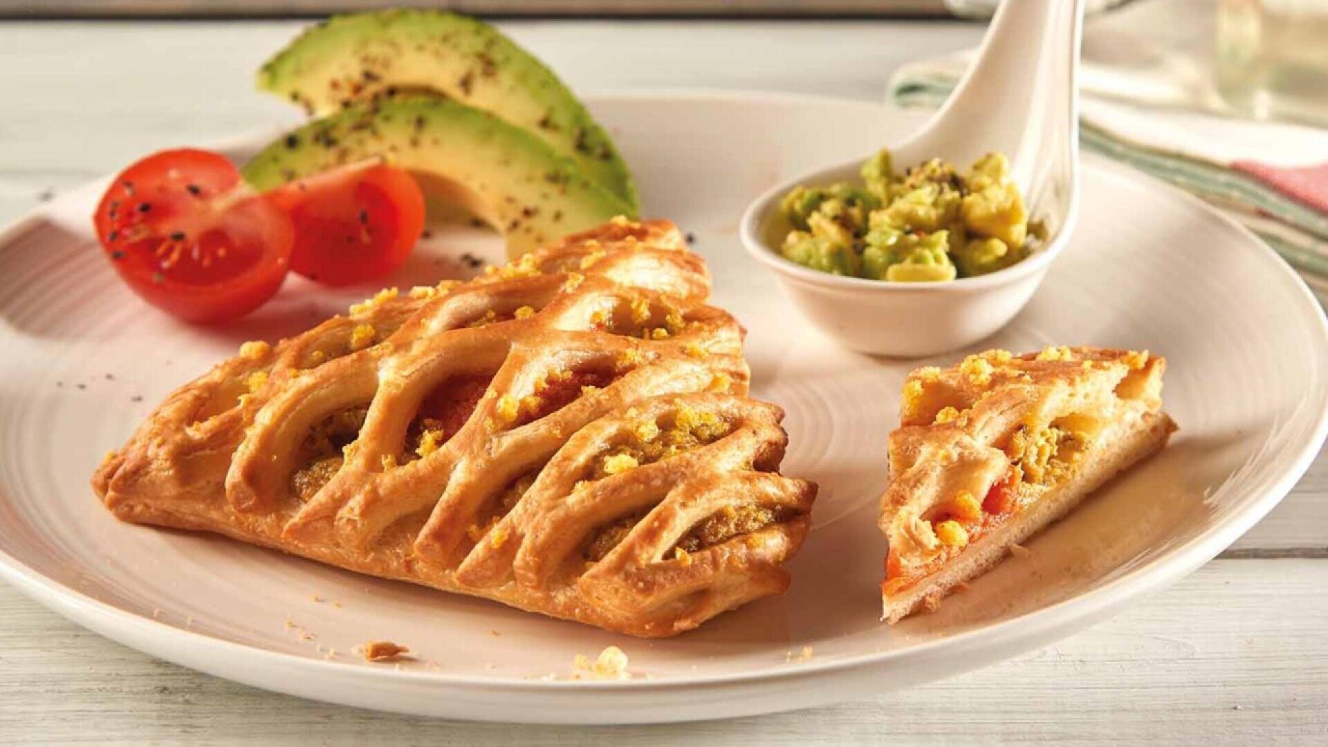 A flaky lattice pastry filled with a savory mixture is served on a plate with sliced avocado, cherry tomato halves, and a small spoonful of chunky dip. One slice of the pastry is cut and set beside the main piece.