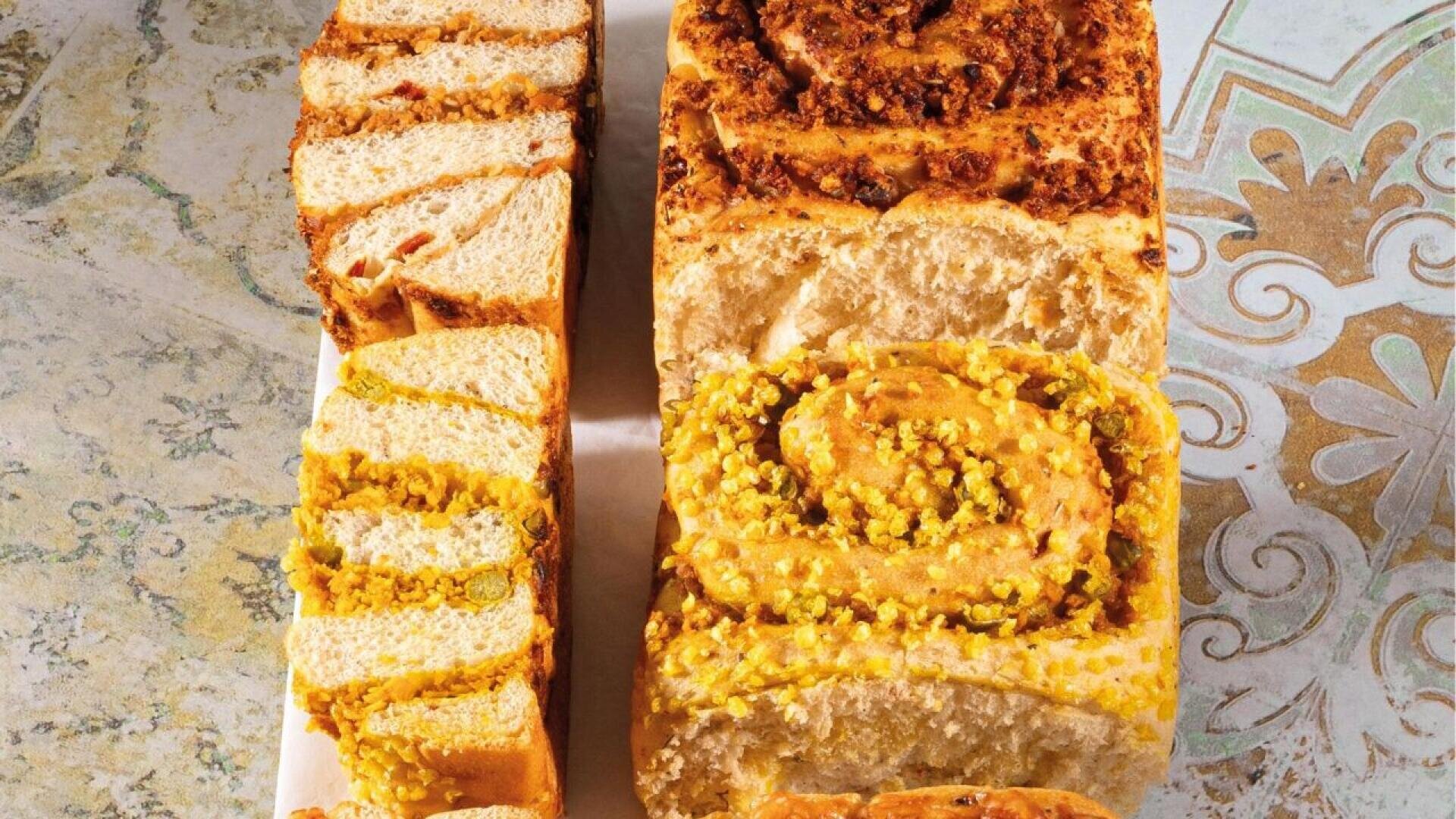 Two loaves of swirled bread, one with a yellow filling and the other with a brown, nutty filling, are sliced to show their textured interiors. They are displayed on a patterned surface.