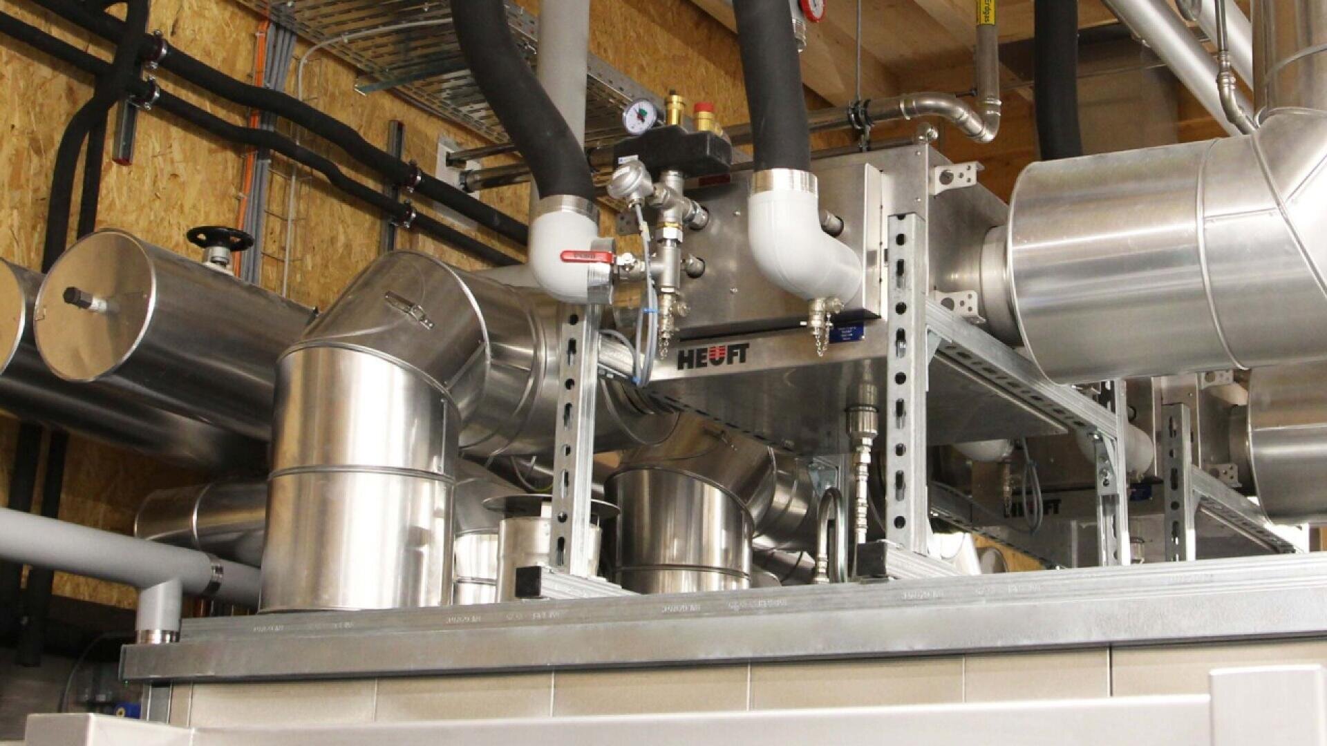 An industrial setup featuring large stainless steel pipes, valves, and insulated ducts interconnected on a metal framework inside a facility with wood-paneled walls and exposed ceiling elements.
