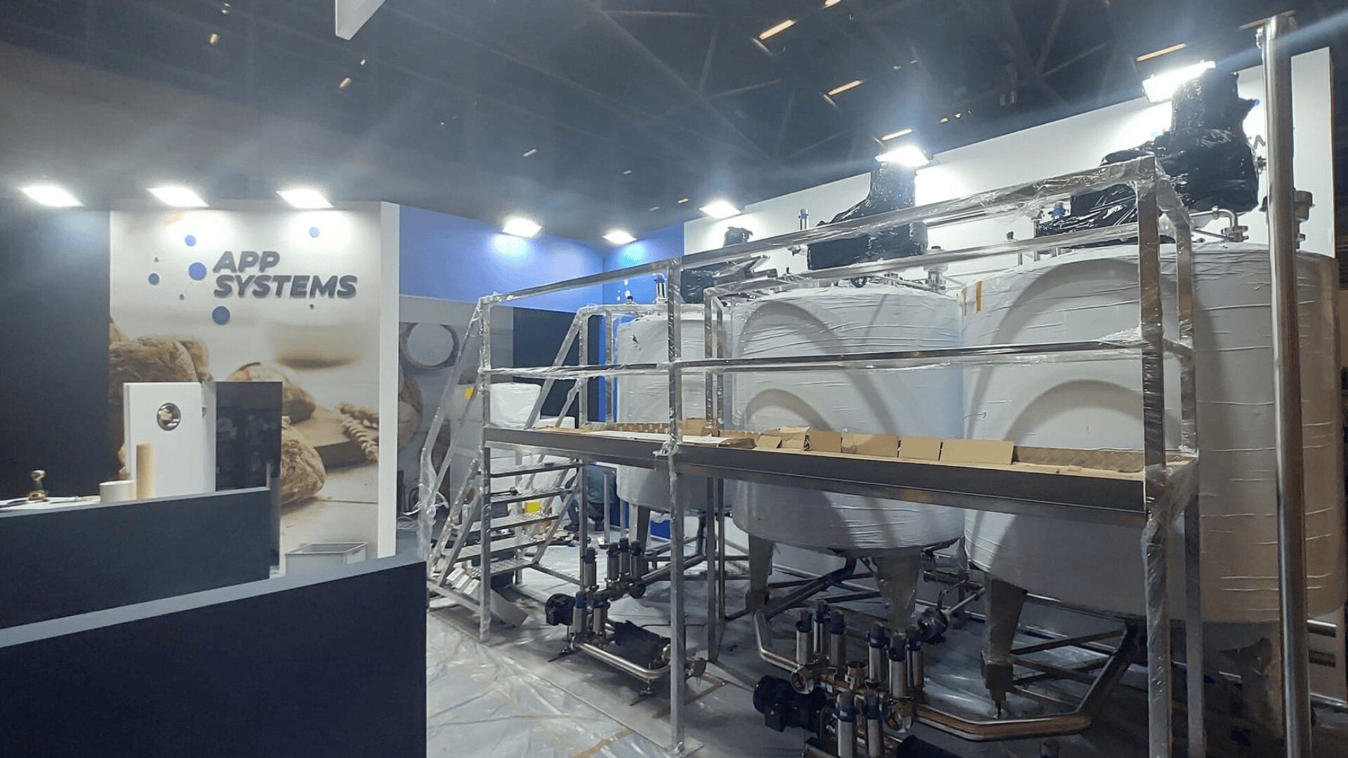 A trade show booth displays large, industrial white tanks and stainless steel piping equipment. The backdrop features the APP SYSTEMS logo with images of grains. Bright lights illuminate the modern setup.