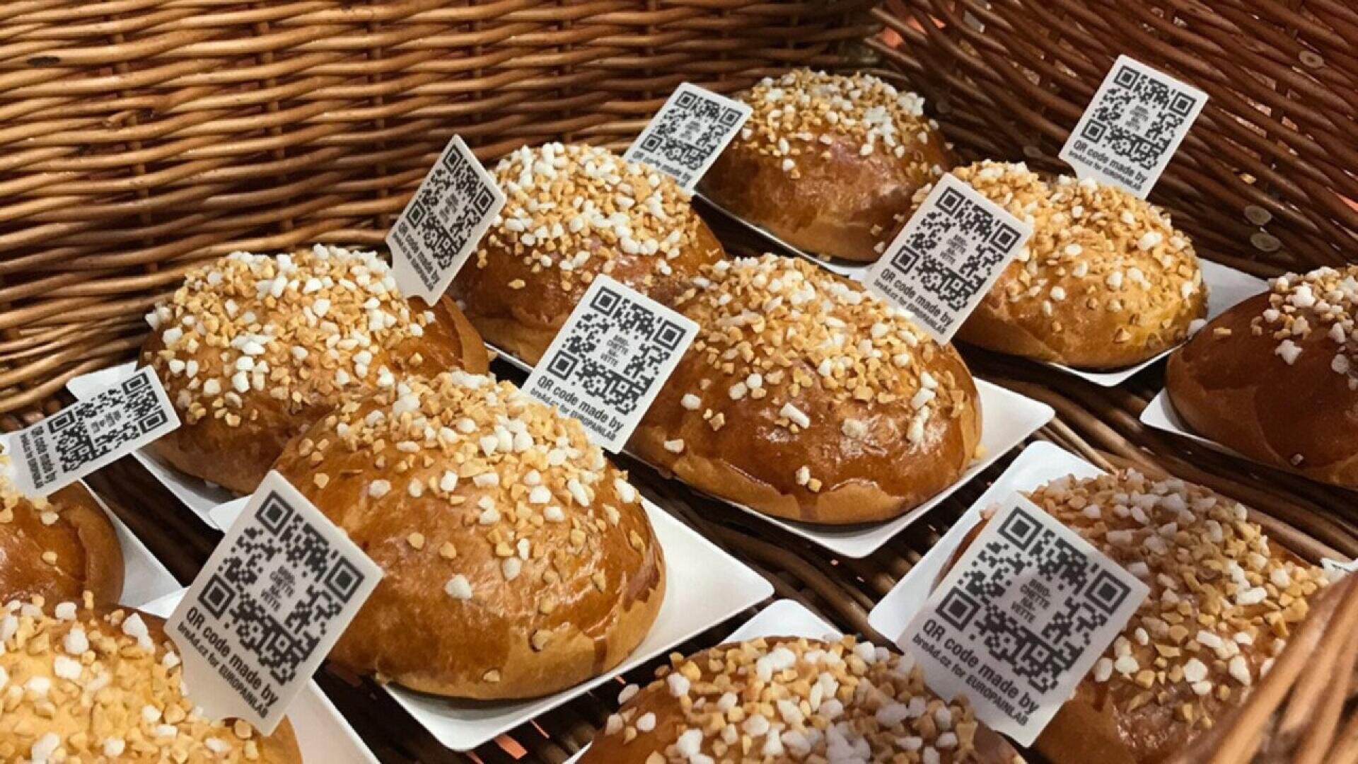 Rundes Gebäck mit Zuckerkristallen auf der Oberseite ist in einem Weidenkorb angeordnet, jedes auf einem kleinen Tablett und mit einem QR-Code-Etikett versehen.