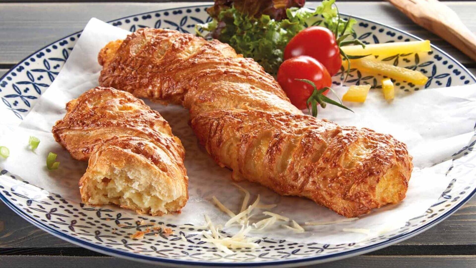 A golden-brown, flaky pastry with a cheesy crust rests on a patterned plate; one piece is broken open to show the soft interior. Garnished with lettuce, cherry tomatoes, and sliced yellow peppers on the side.