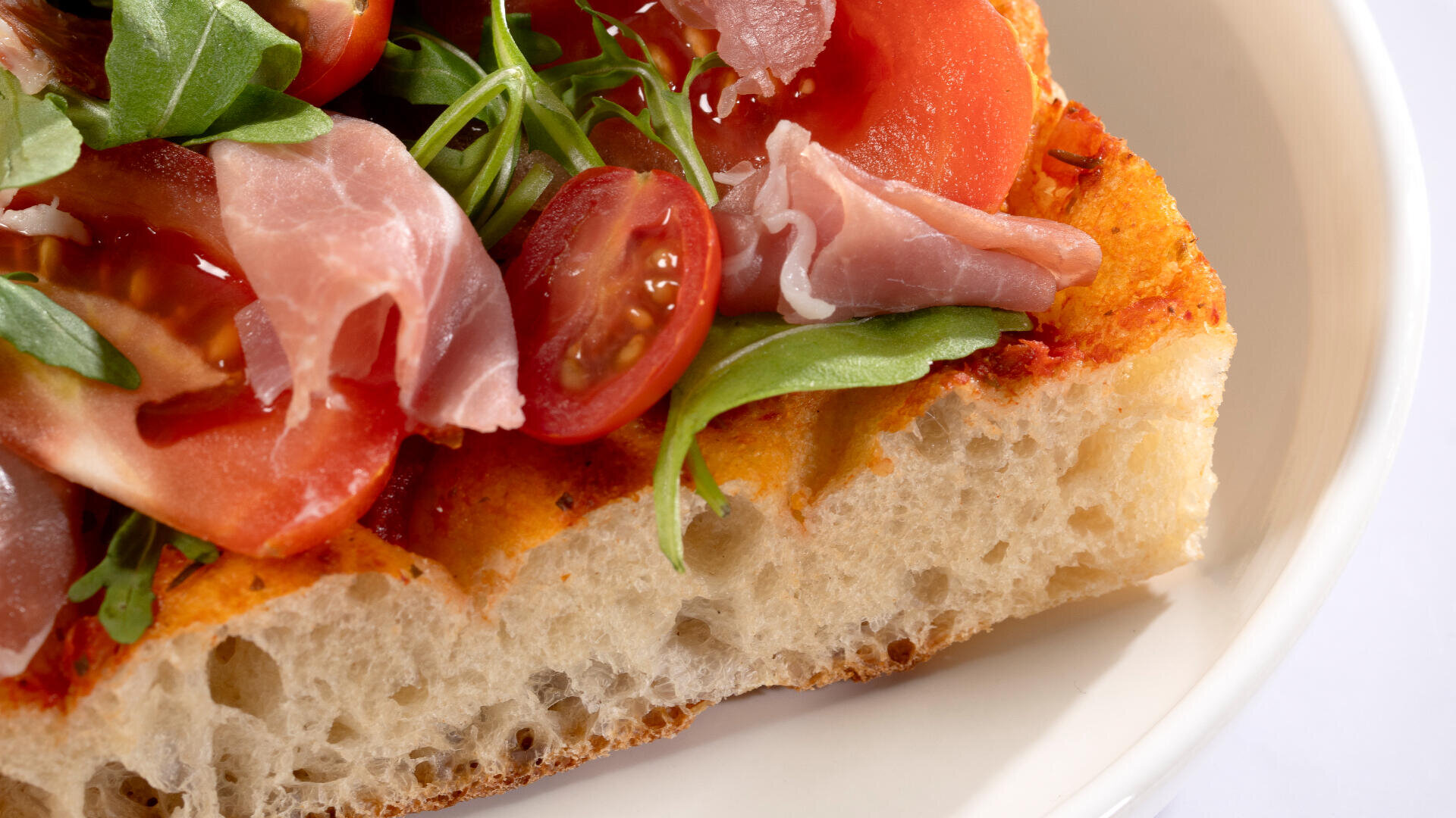 Eine Nahaufnahme einer Scheibe Focaccia-Brot, belegt mit Rucola, Kirschtomaten, Prosciutto und Tomatenscheiben auf einem weißen Teller.