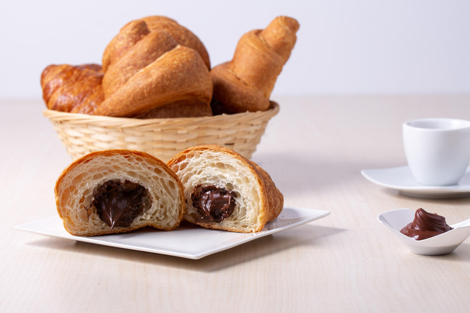 A plate with a halved croissant filled with chocolate cream, a spoonful of chocolate spread, a basket of croissants, and a white espresso cup in the background on a light wooden table.