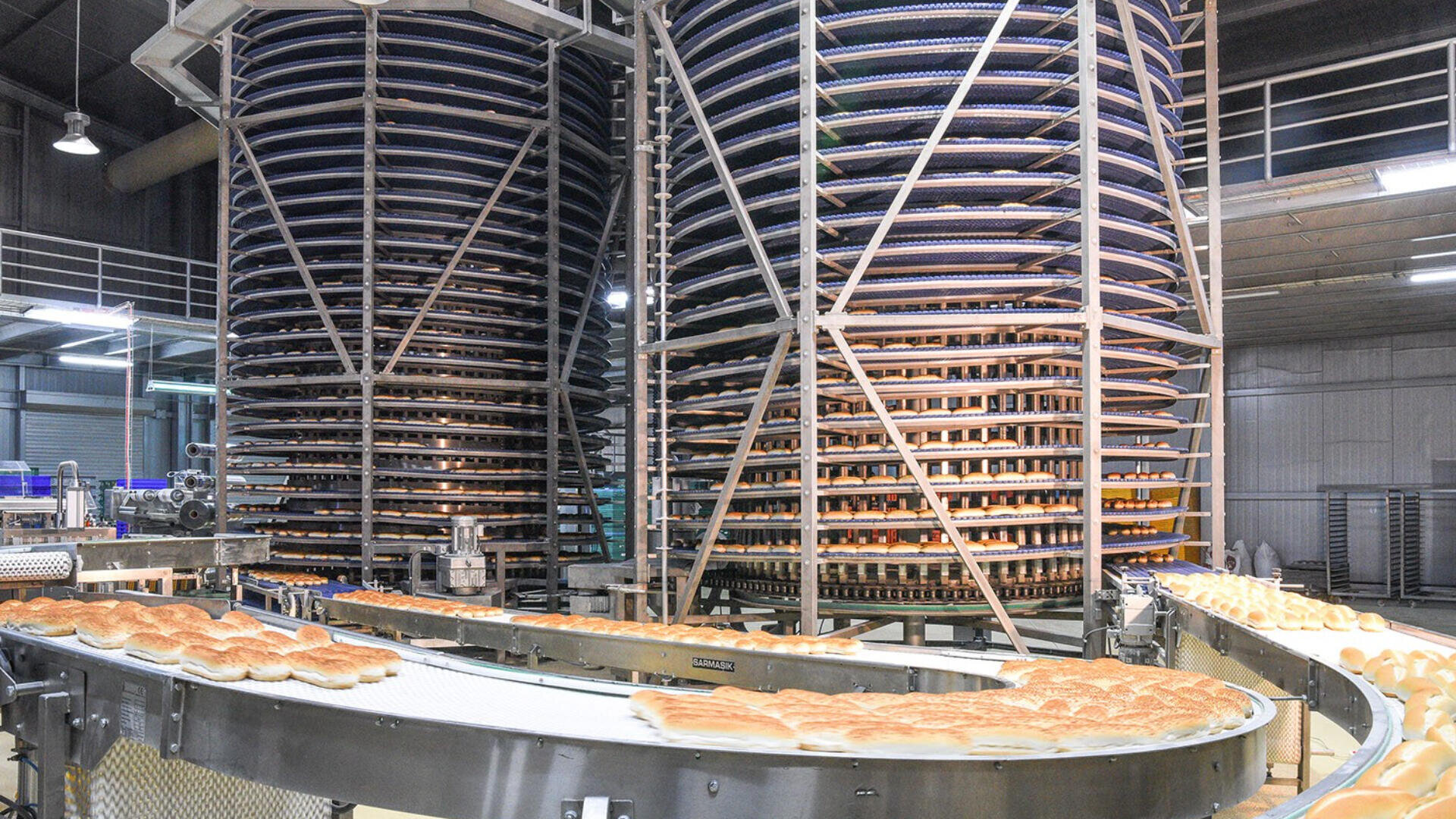Große automatisierte Bäckereifertigungsstraße mit Förderbändern, die Reihen von Brotlaiben transportieren, und hohen spiralförmigen Kühltürmen, die mit Brot gefüllt sind, in einer Industrieanlage.