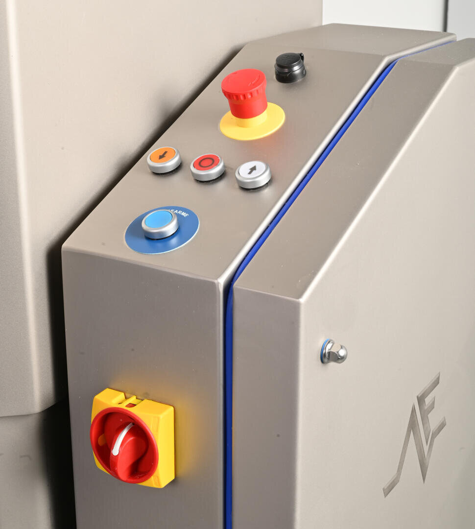 Close-up of a metallic machine control panel with various buttons, including a large red emergency stop button, a blue button, three smaller buttons, and a yellow and red rotary switch. The panel has a modern, industrial design.