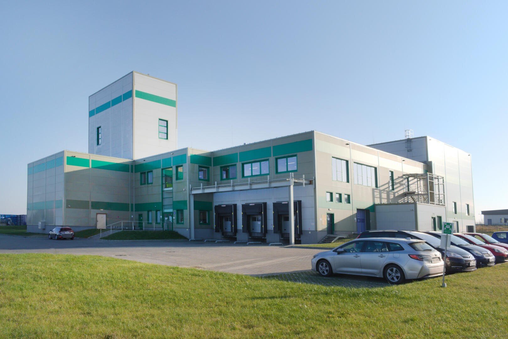 A modern industrial building with green and white exterior panels, several loading docks, and a few parked cars on a driveway, set against a clear blue sky and grassy foreground.