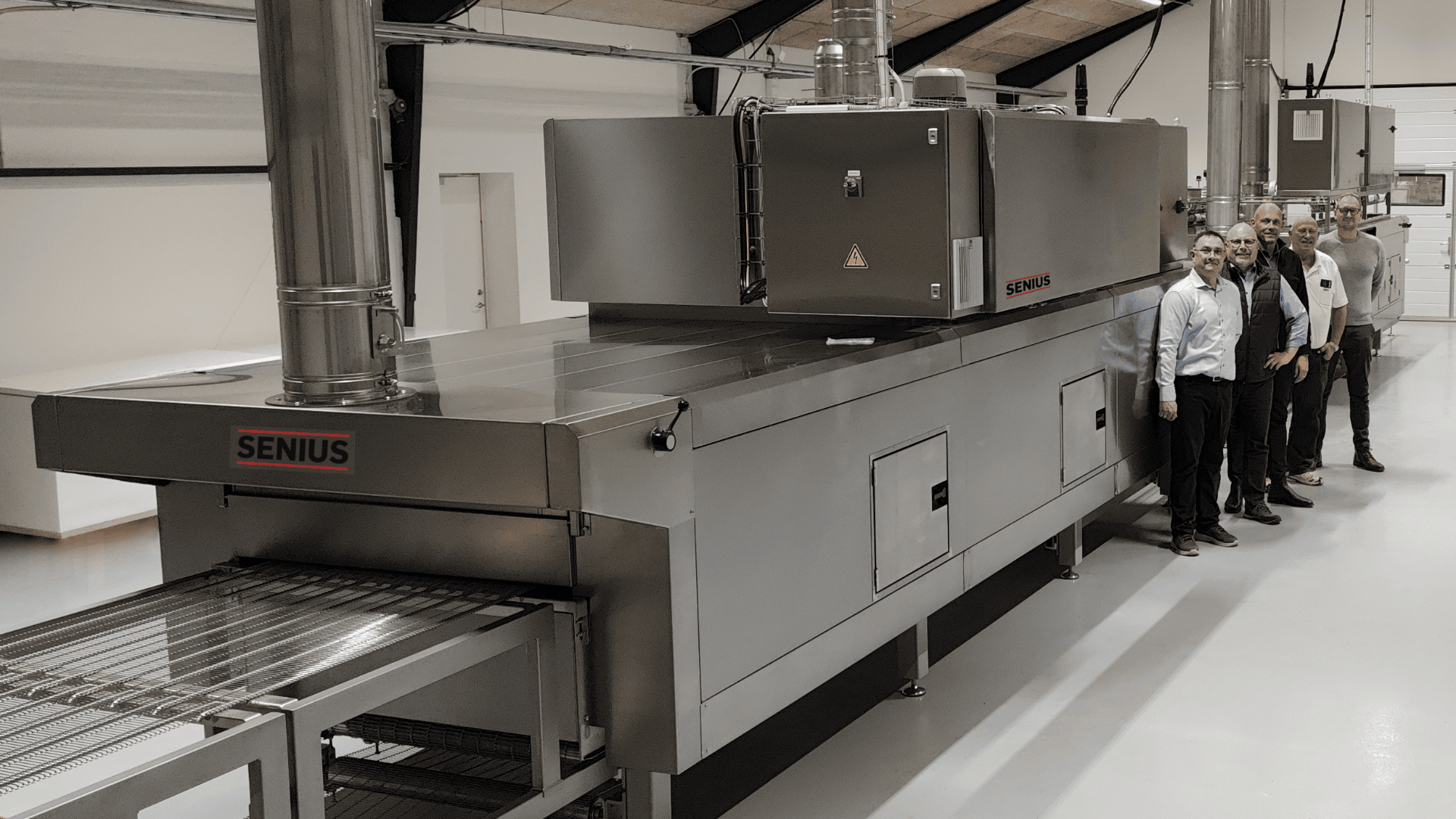 Fünf Männer in Geschäftskleidung stehen neben einem großen industriellen Senius-Ofen in einer sauberen, modernen Anlage. Der Ofen verfügt über Metallflächen, Lüftungsöffnungen und ein Förderband.