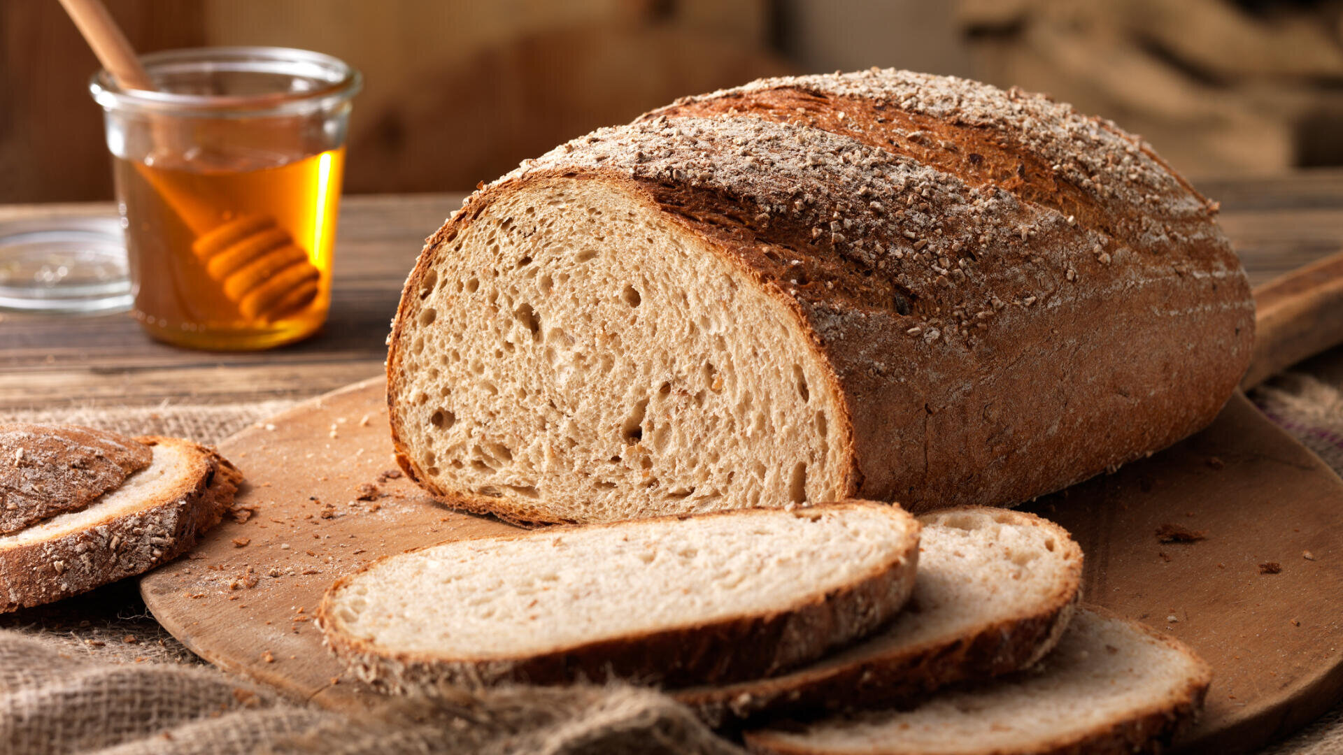 Ein Laib rustikales Vollkornbrot liegt auf einem Holzschneidebrett, einige Scheiben sind bereits angeschnitten. Im Hintergrund ist ein Honigglas mit Honiglöffel zu sehen. Die Atmosphäre ist gemütlich und rustikal.