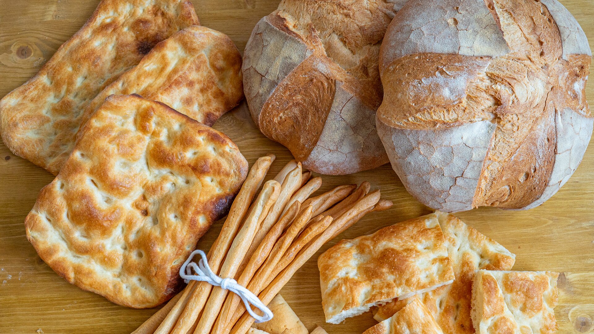 An assortment of bread on a wooden surface, including round loaves with a cross pattern, flat rectangular focaccia, crispy breadsticks tied together, and sliced pieces of golden flatbread.