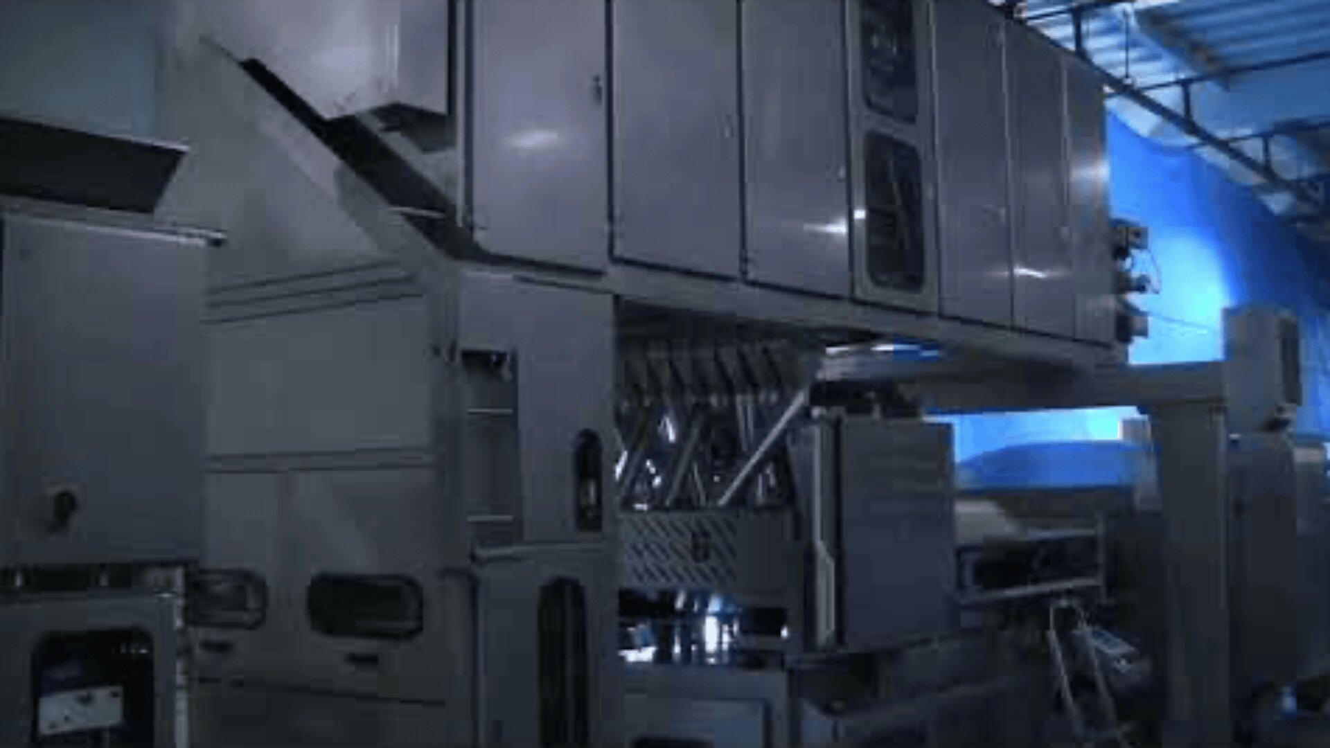 Große Industriemaschine mit Metallplatten, Entlüftungsöffnungen und Rohren in einer Fabrikumgebung, beleuchtet von Oberlichtern.