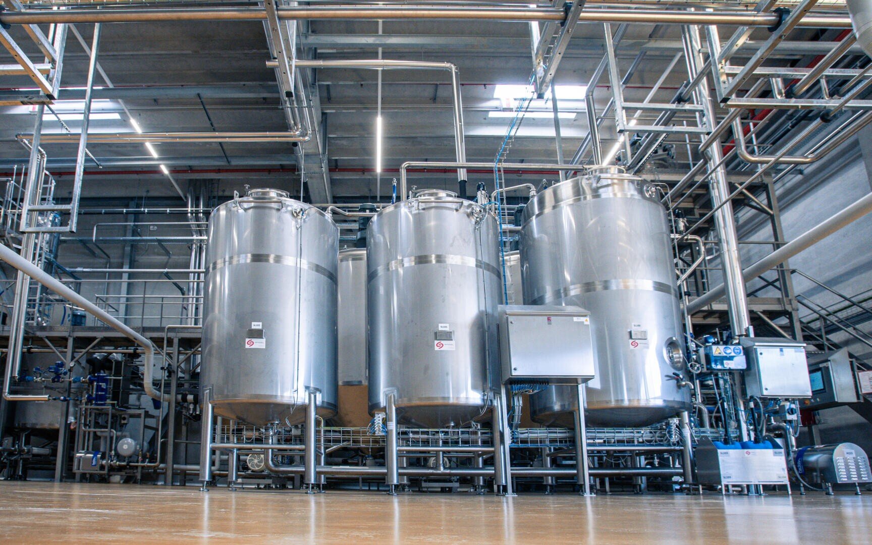 Large stainless steel tanks and interconnected pipes inside an industrial facility, likely used for processing or storing liquids such as in a brewery or food manufacturing plant.