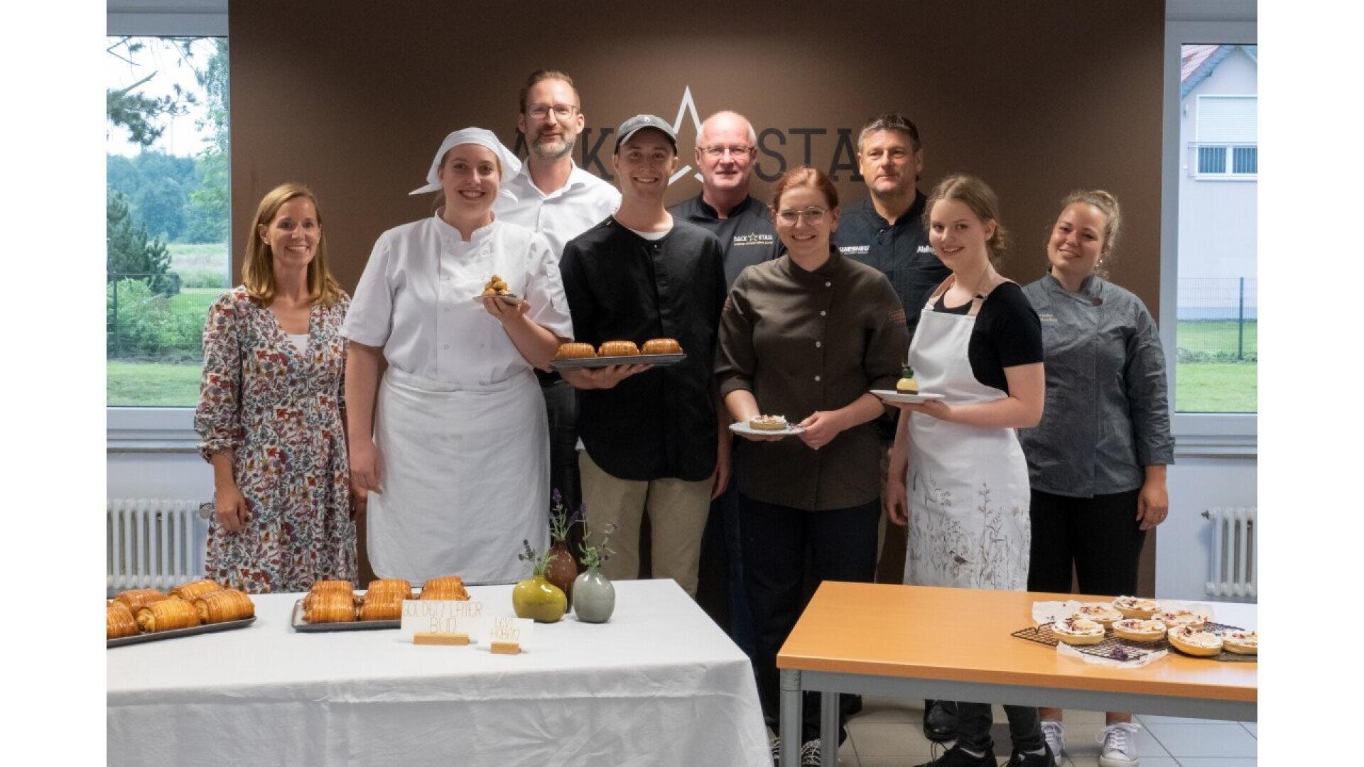 A group of ten people, including chefs and staff, stand smiling behind tables with baked goods in a bright room. Some hold plates or trays of pastries, and everyone looks happy and proud of their work.